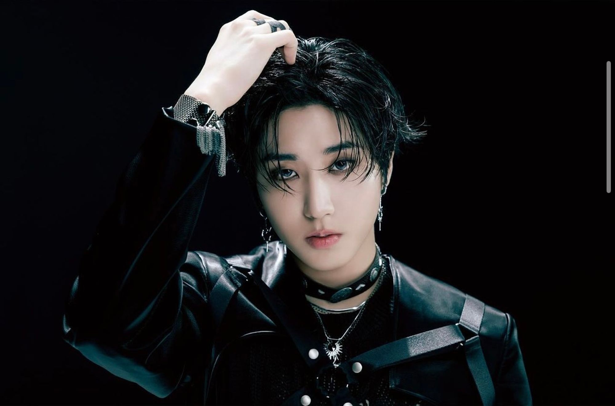 Stray Kids member HAN from the chest up wearing a black jacket and leather harness, one hand touching the top of his head as he looks directly to the camera.