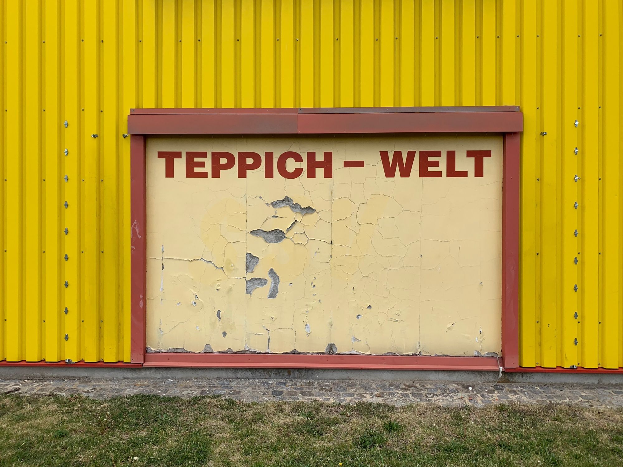 Fensterlose gelbe Industriehalle aus Metallprofilen. In der Mitte eine leicht lädierte Plakatwand mit der Aufschrift „Teppich-Welt“.