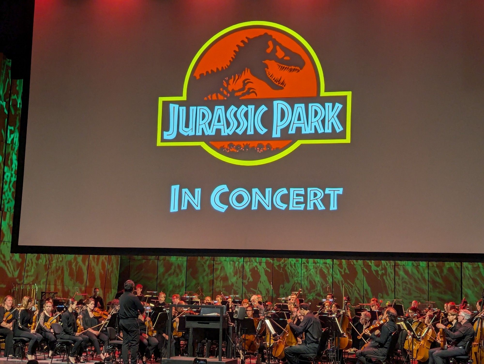 The Jurassic Park logo projected on a huge screen, under which reads 'in concert' in the jpark font. Under the screen you can see a full orchestra seated.