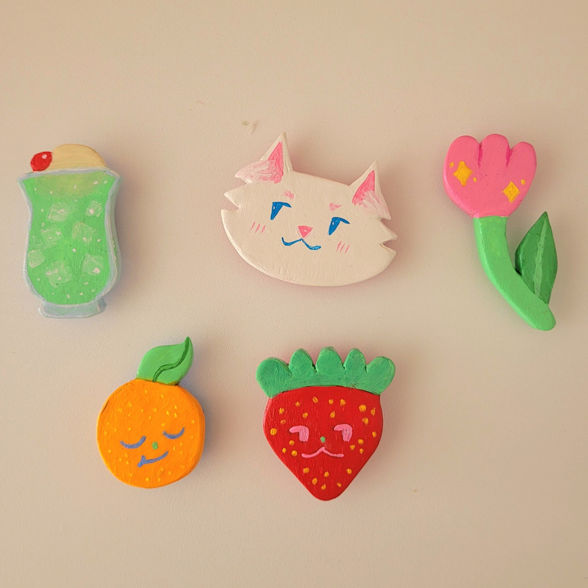 A set of five colorful handmade clay magnets on a white surface. One is a melon cream soda float with green soda and ice cubes in a glass, topped with vanilla ice cream and a cherry. One is the head of a fluffy white cat with a mischievous expression. One is a pink tulip flower with a green stem and leaf. One is an orange fruit with a leaf and a sleepy smiling expression. One is a red strawberry with a lazy smiling expression. 