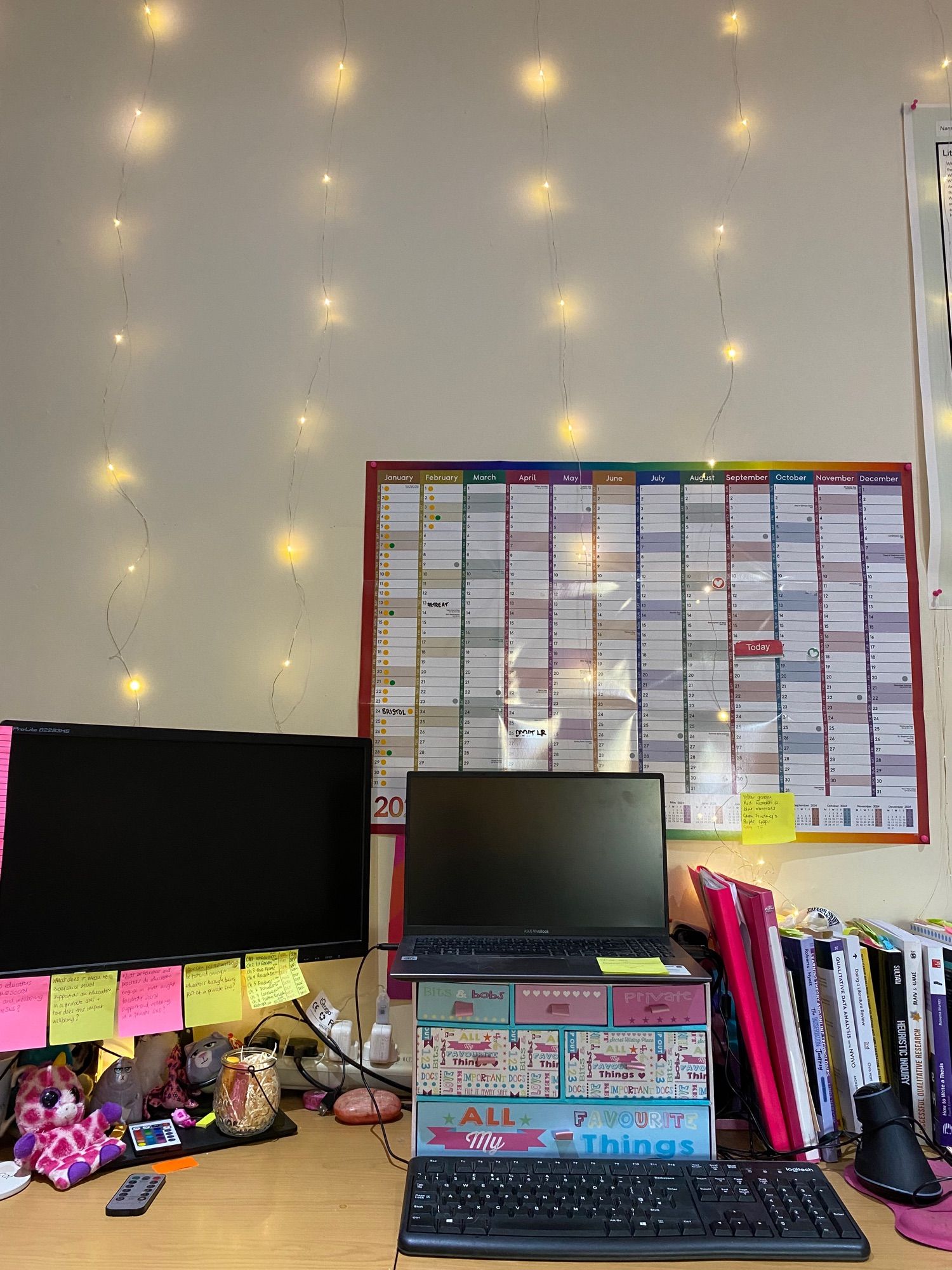 My desk with computer and wall-calendar complete with fairy lights,  messy sticky notes, books and a few fluffy toys