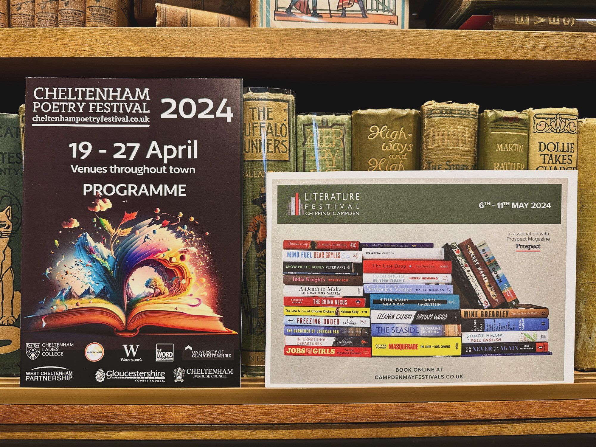 Two literary festival programmes side by side on a brown bookcase with decorative cloth antiquarian books behind