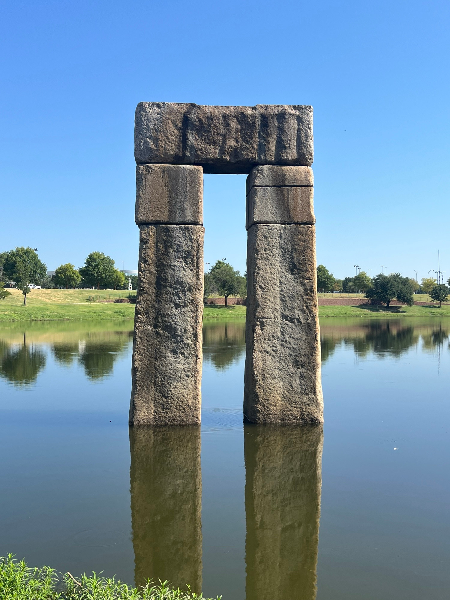 A stone arch way made of two vertical rectangular pillars, topped by stone cubes, then connected by a cross over. Opening up from within and towards the lake. The Gateway of Lies. 