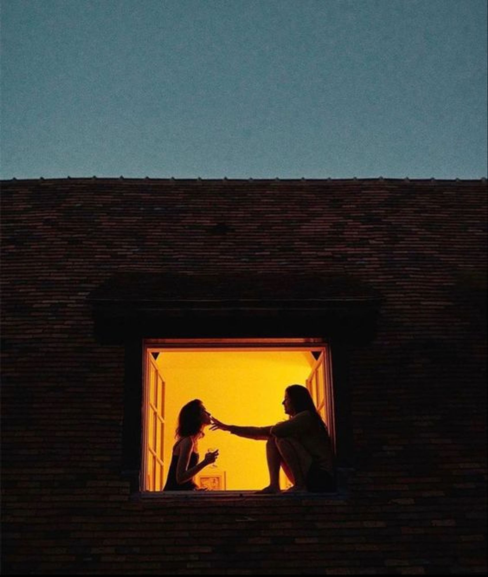 Photo of two women sitting in an open window, silhouetted by a golden glow from inside. The window is framed by the darkened roof of the building, and the top third of the image is a sky darkening as if at twilight.