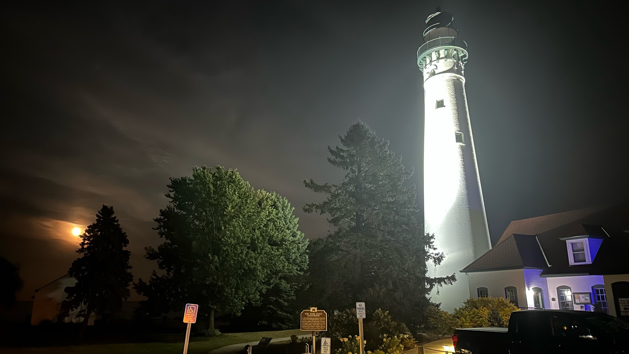 Super moon peaking out from behind a spruce tree at left of frame. More trees in kind frame. Lake Michigan lighthouse in front right of frame. Dark skies with some wispy stratus clouds.