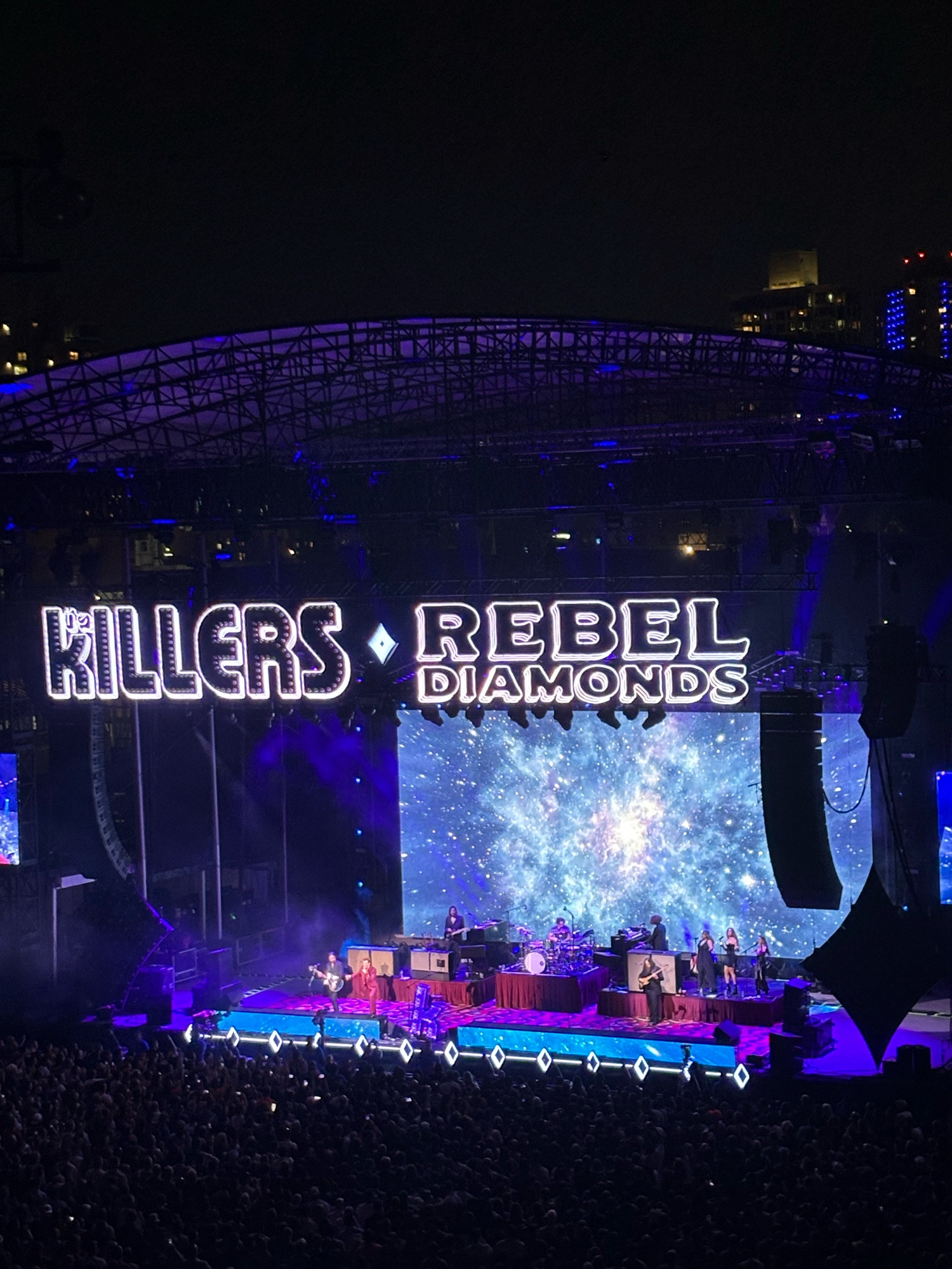 Photo of the band The Killers on stage with space graphics, Killers Rebel Diamonds in blue lighting above