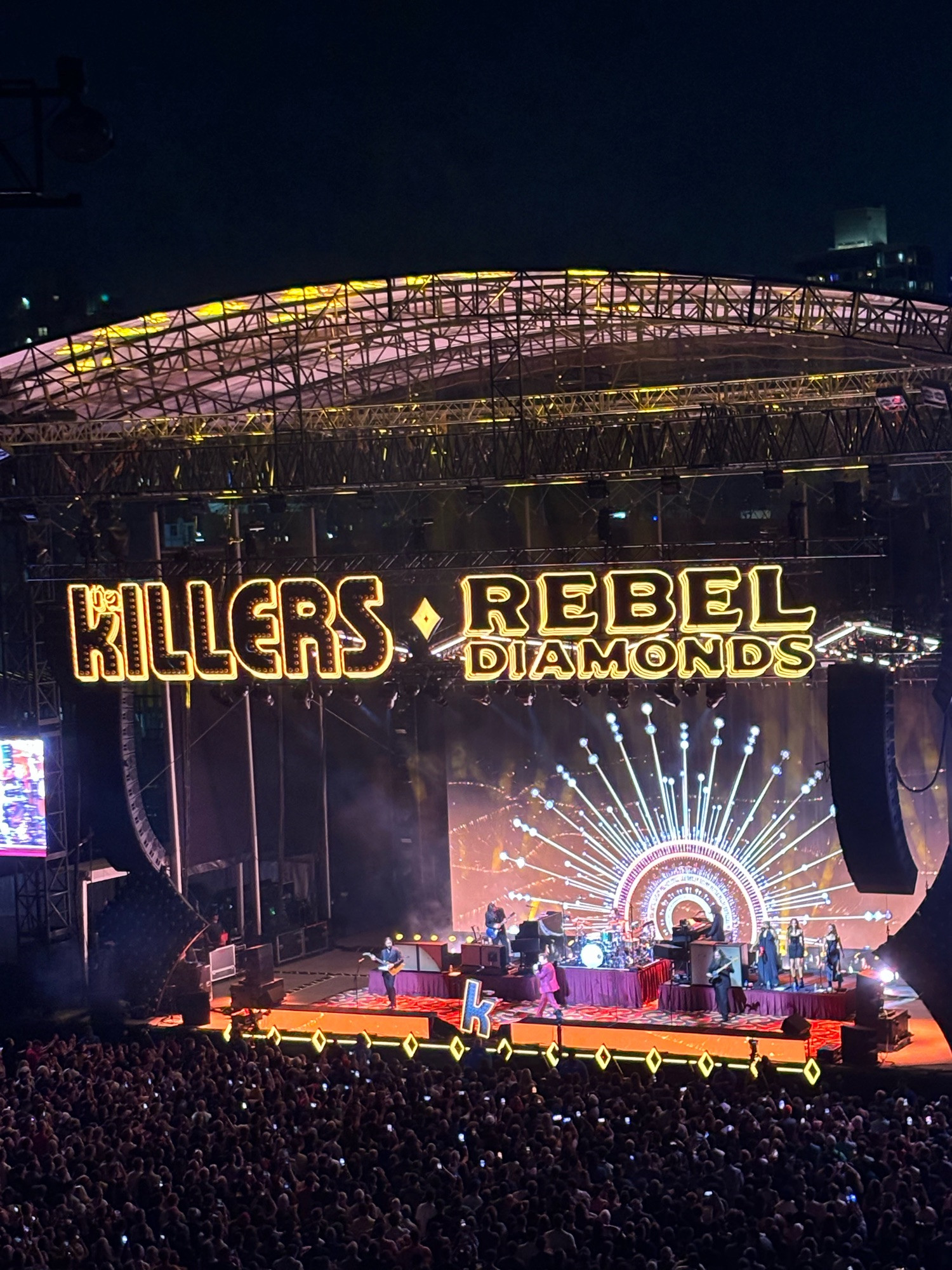 Photo of the band The Killers on stage with Killers Rebel Diamonds in yellow lighting above