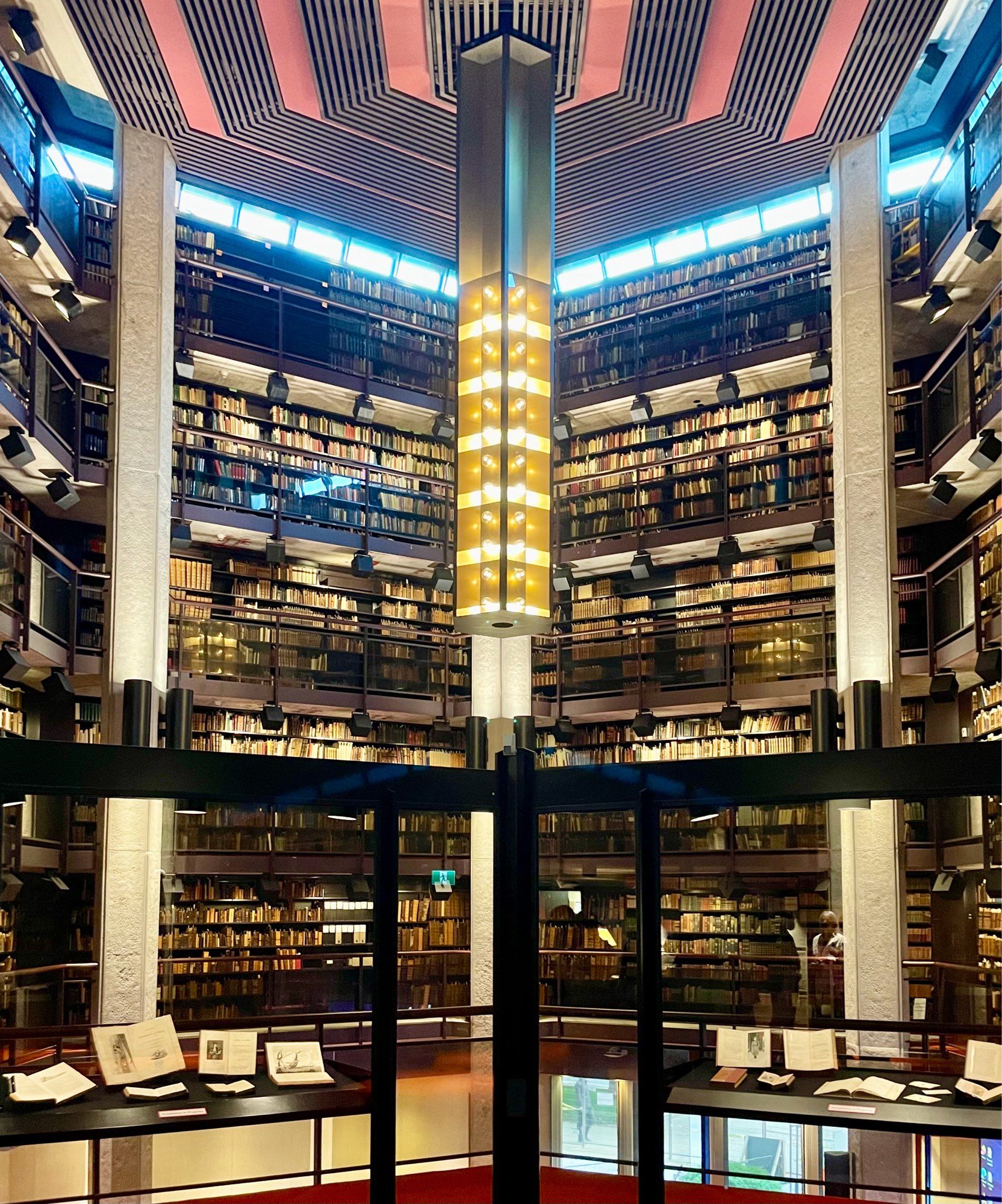 inside an octogonal concrete brutalist building walled full of book stacks and a large art deco lamp in the middle.