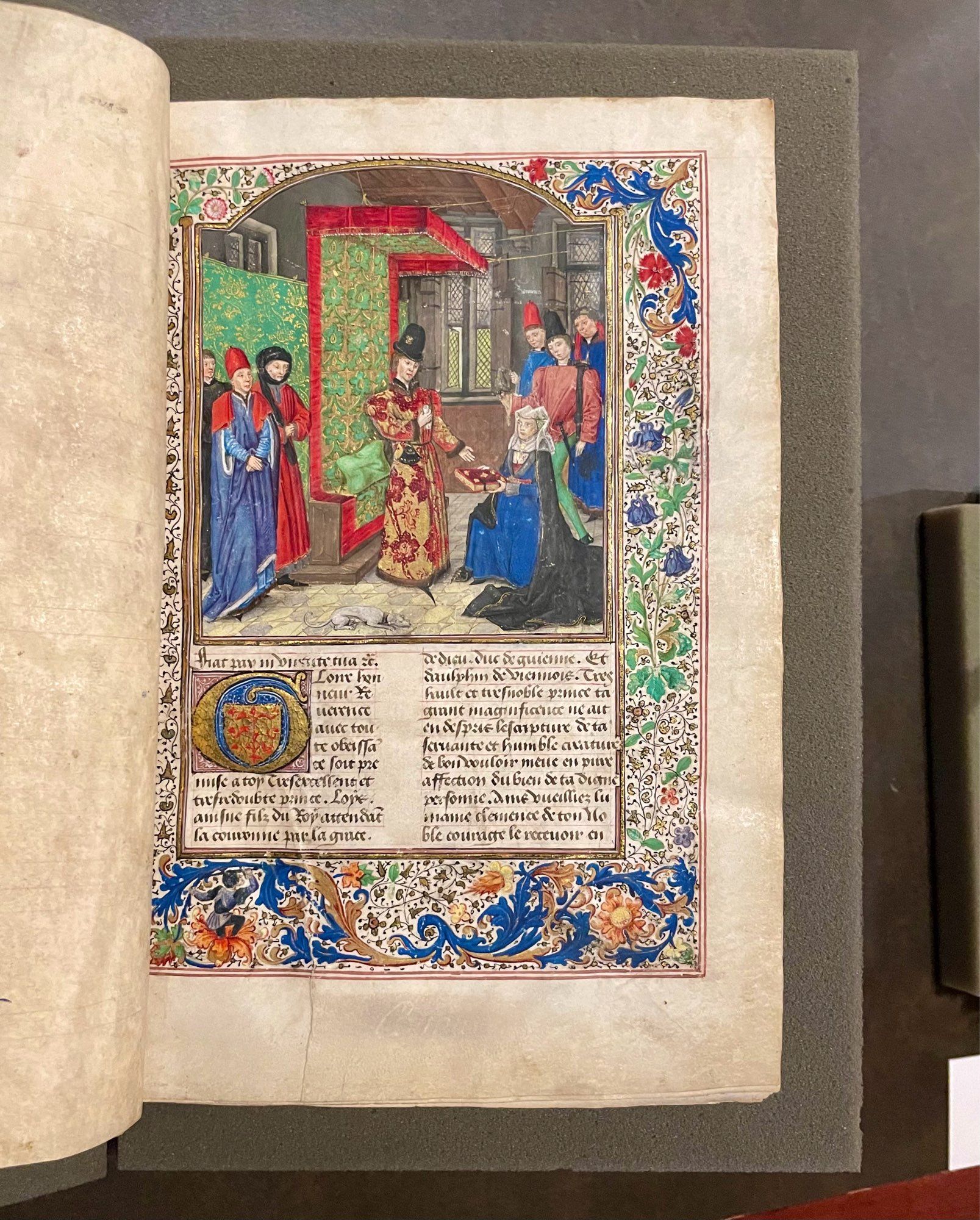 medieval manuscript page with text, flower decoration and a large miniature showing a kneeling Christine de Pizan offering her book to a prince. Fancily dressed male attendance in the background