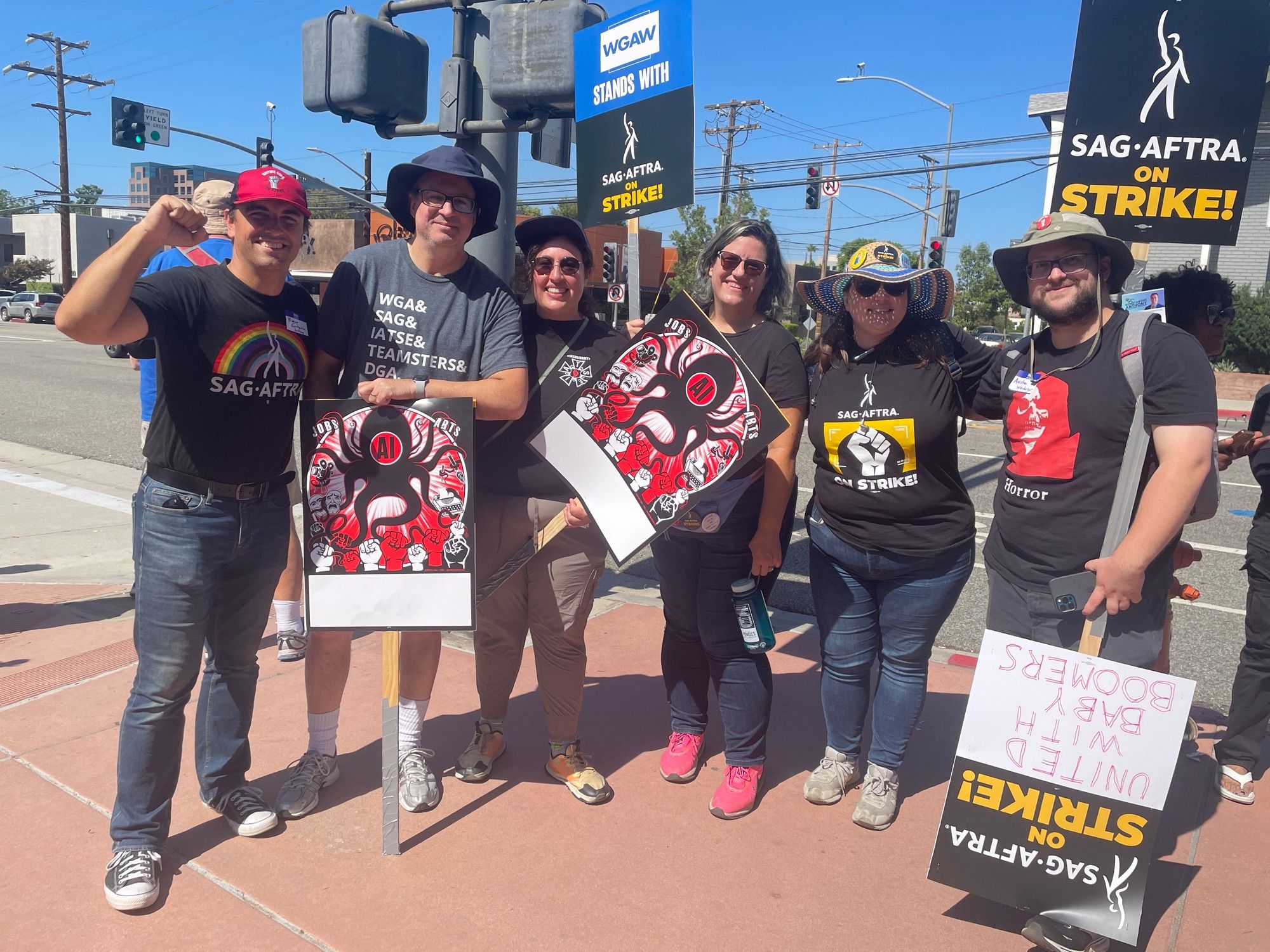 Six people with picket signs, wearing SAG-AFTRA,  IATSE and other union shirts