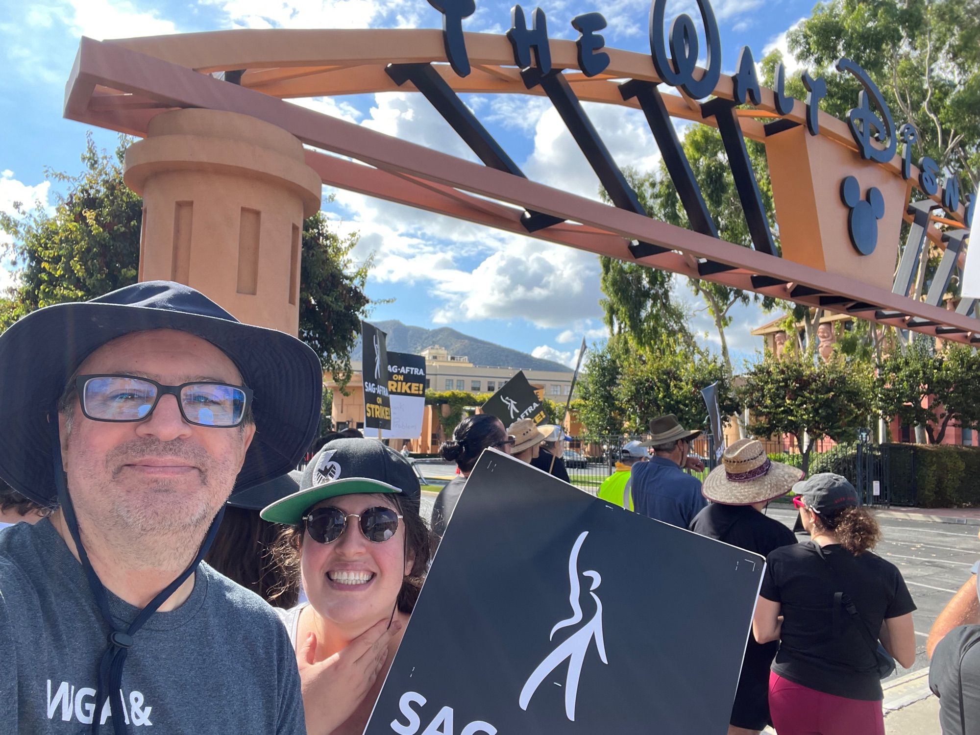 A man and a woman standing in front of the main Walt Disney gate in Burbank, holding a SAG-AFTRA strike sign, surrounded by other picketers.