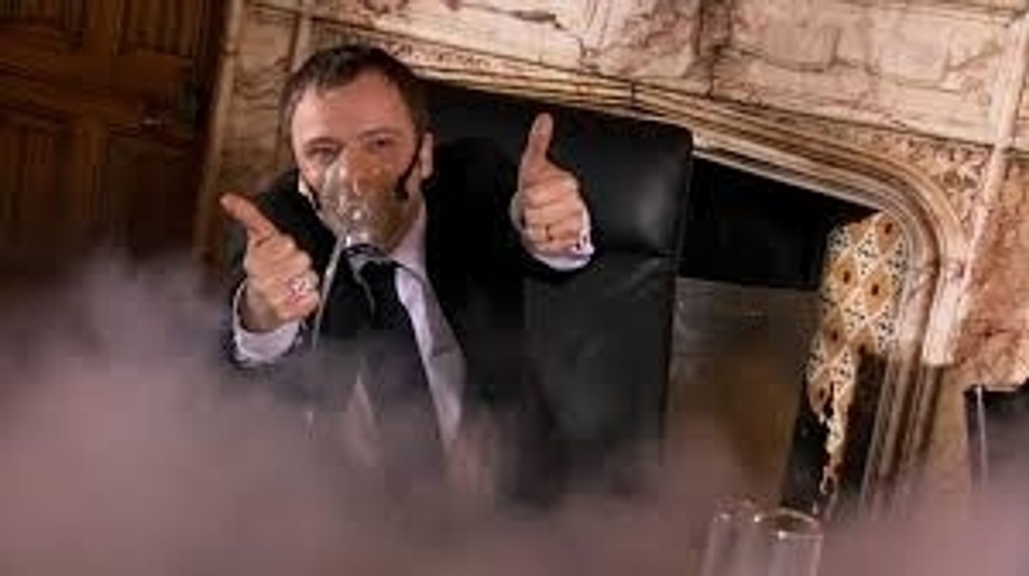 John Simms as the Master in Doctor Who. He's sitting in front of a marble fireplace while wearing a suit and a gas mask and giving a gleeful thumbs up to the camera as gas fills the room around him.