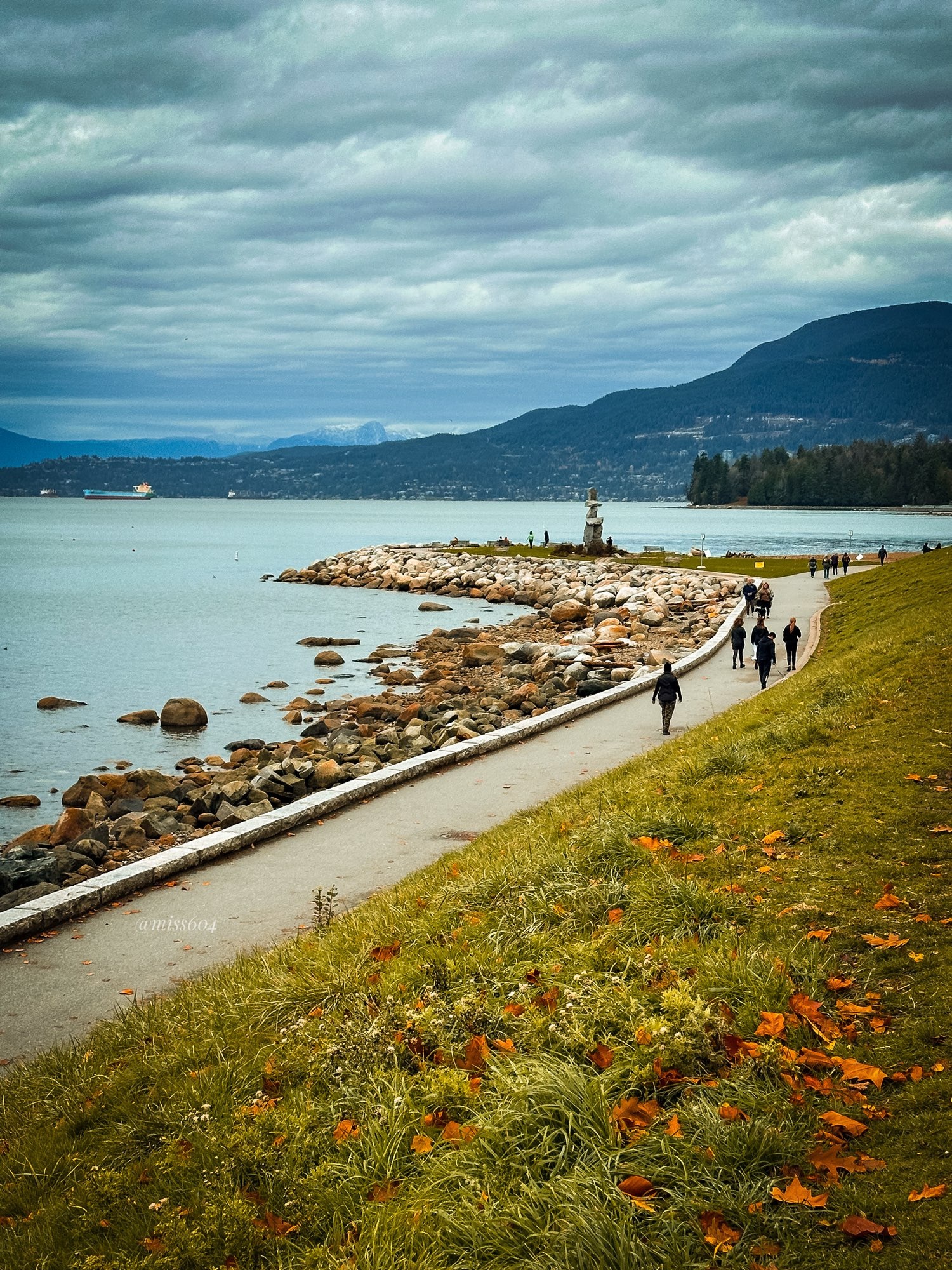 A grassy hill on the right, the paved Seawall path through the middle of the shot, and English Bay on the left. Leaves scattered across the grass, people walking on the Seawall. The sky is cloudy but the north shore mountains loom in the background