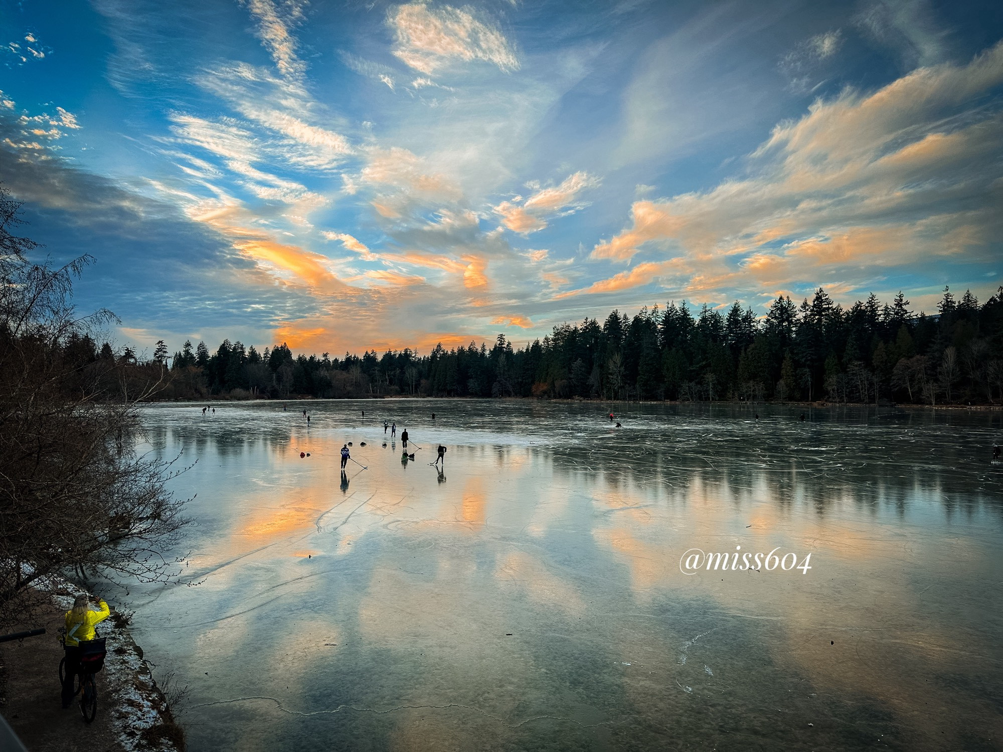 A person in a bright jacket takes a photo of the lagoon from a path on the shore. On the lagoon, the surface is frozen and people play hockey. The sunset is reflected on the surface