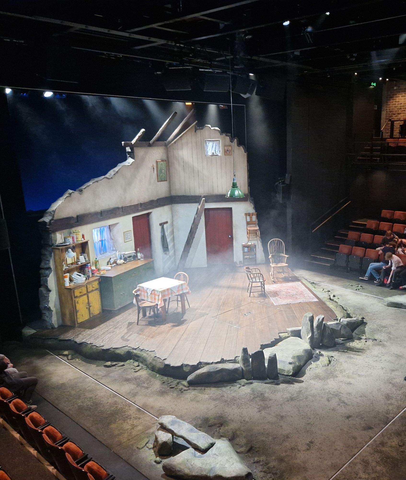 Stage set of Lieutenant of Inishmore. A cut-away section of an old Irish cottage with kitchen and seating area. Around the cottage are small boulders.