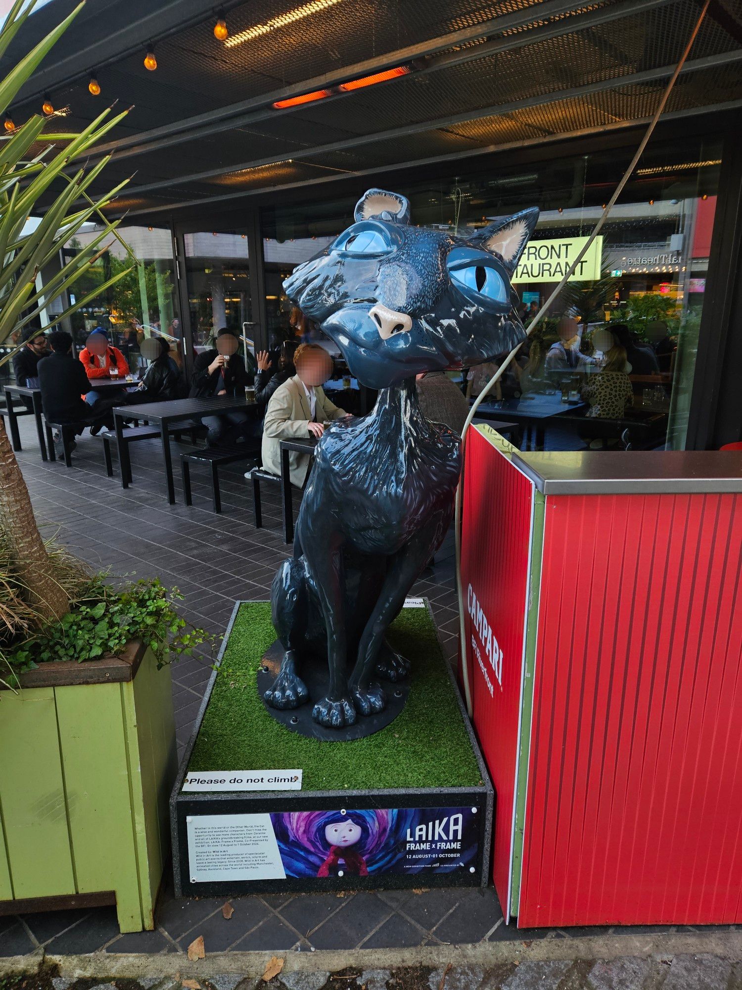 At the side of a fancy cinema building in London is a large model of the cat from the film "Coraline" sat up and looking to his left. The model is on a stand with fake grass below the model itself and behind it are people sat down in tables in the outdoor area of a fancy cinema's bar (BFI Southbank, London, UK).