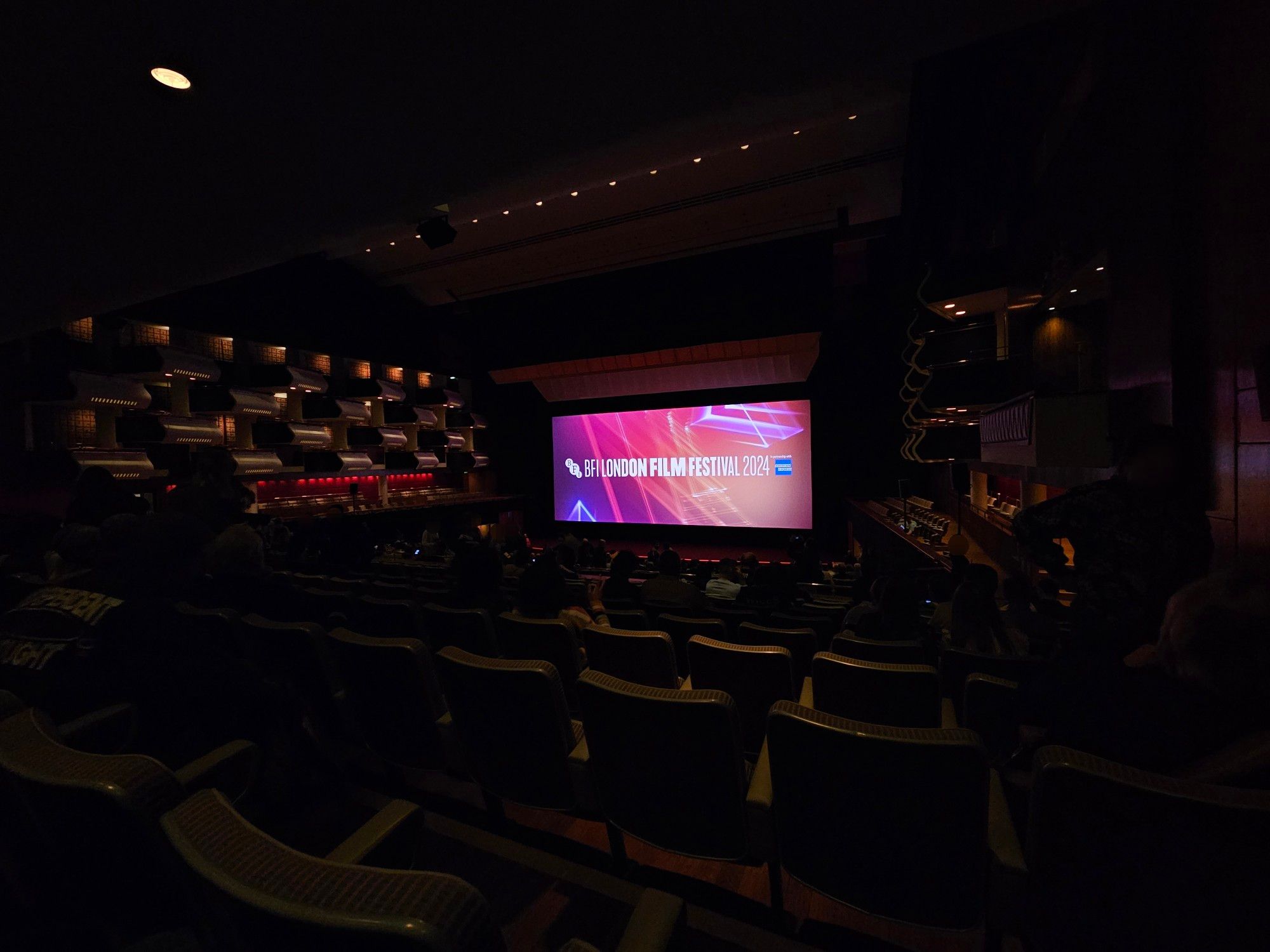 Inside a huge auditorium that has its lights on while people slowly come in and sit in its many seats. An artistically laid-out array of speakers and VIP seats adorn the sides of the view, while at the centre is a huge makeshift screen in a "Cinemascope"-style shape displaying a static screen that adorns the logo for the BFI London Film Festival 2024, alongside the logo for American Express (which gets an "In Assocation With" text above it) and with an abstract background of soft pink, purple and orange hues and abstract angular shapes accompanying it all. The Gala, at this point of the photo, is still about 10-15 minutes from starting.