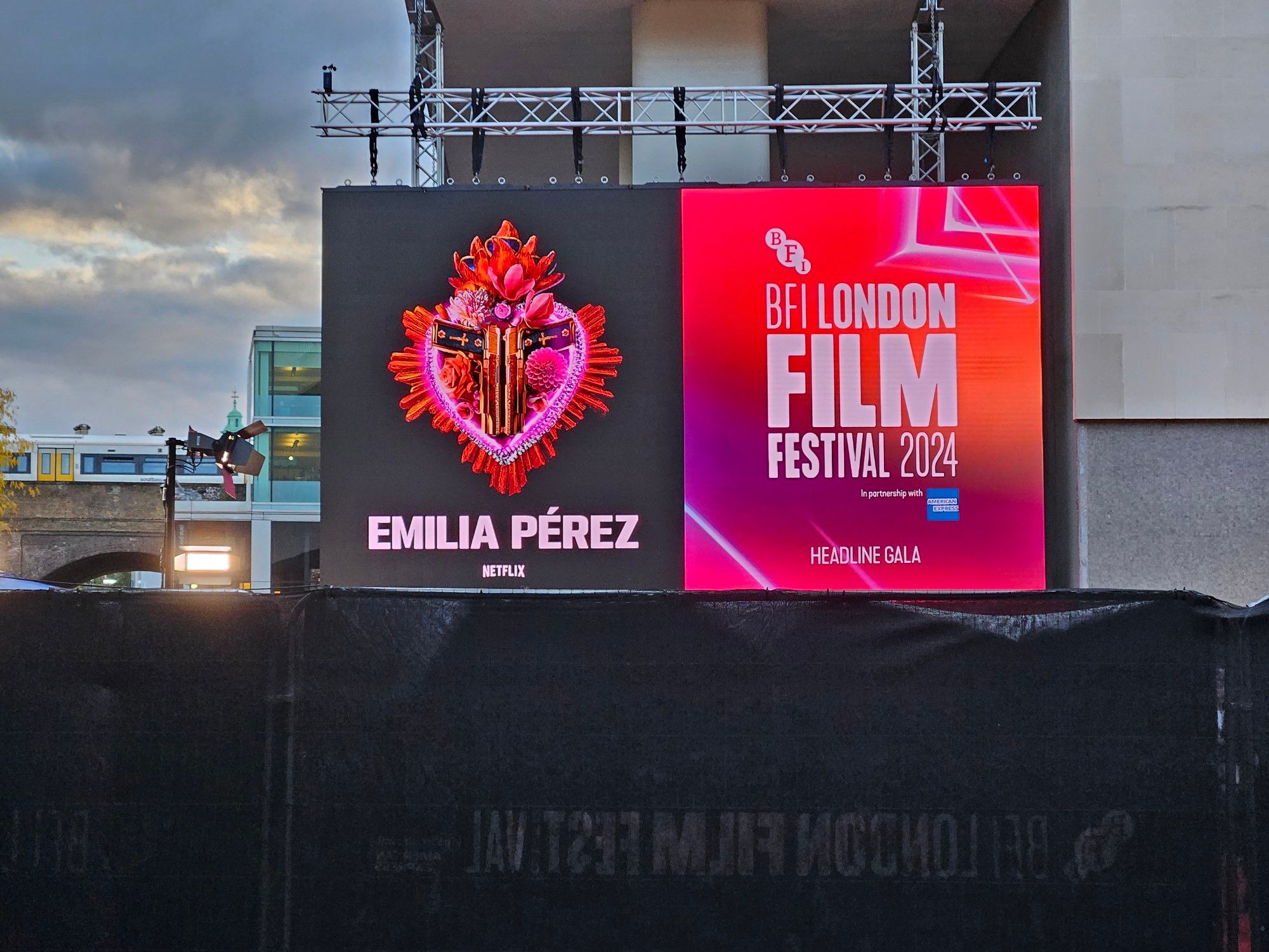 An overcast day in October nearing its sunset point. The side of a large white building dominates the view, with a large sign with electronic screens on it afixed to it. The screens show the poster to the left of a film called "Emilia Perez" (showcasing a stylized heart with flowers and two gold-and-black pistols inside it) and a poster to the right for the BFI London Film Festival 2024 (with hues of pink, purple and orange accompanying abstract angular shapes). The bottom of the London Film Festival poster has text that reads "Headline Gala". In the foreground are gates coated in a black fabric, forming the barriers of a path. This is a view of an entrance to a Gala of that film's Red Carpet, leading to an entrance to its venue, the Southbank Centre (London, UK).