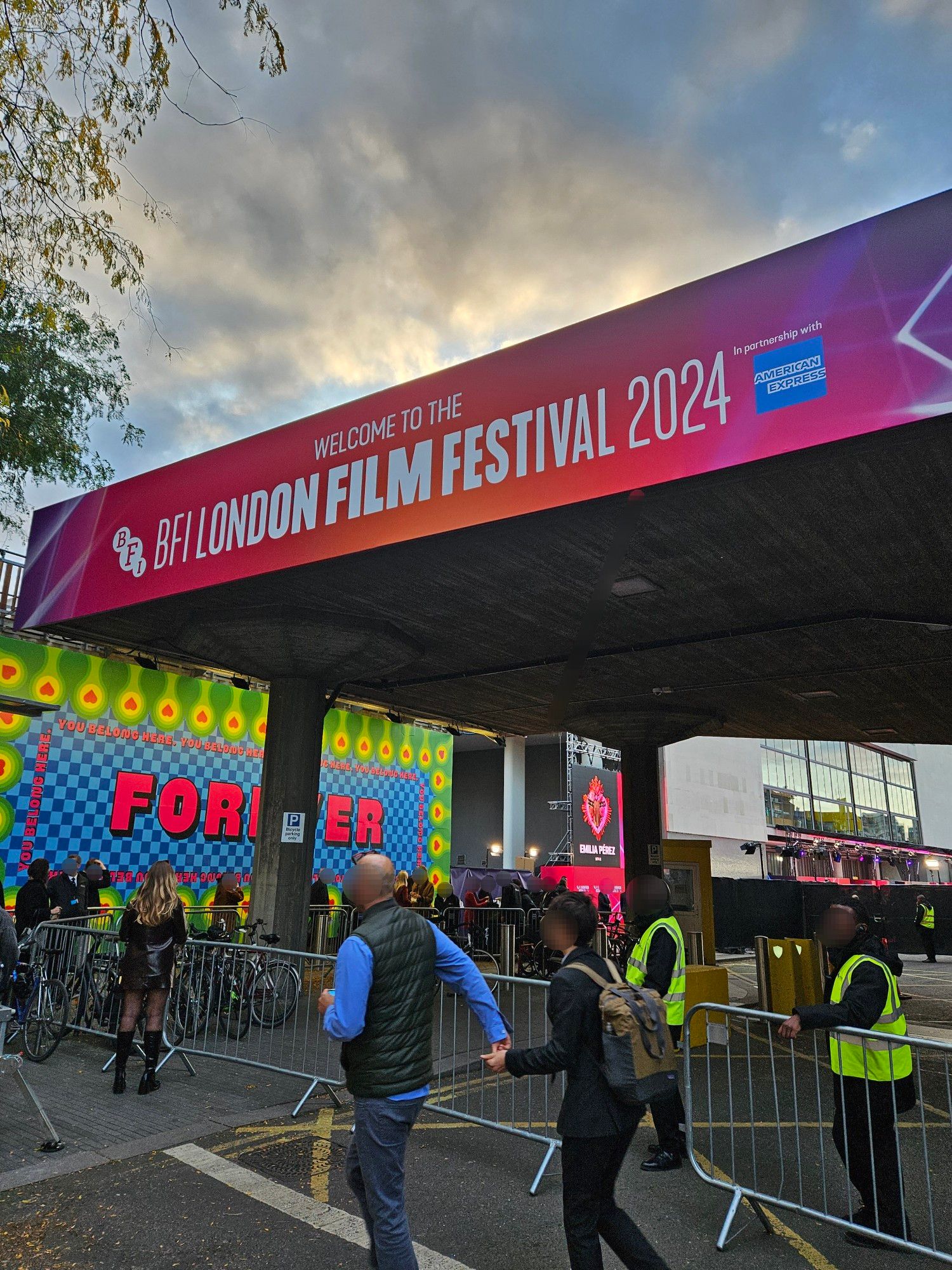 An overcast day nearing a sunset point. Trees with light leaves of green and yellow peek at the top-left of the view, which is dominated by a structure in fr⁸ont of a special entrance into a large white building that adorns a wide overhead sign reading "WELCOME TO THE BFI LONDON FILM FESTIVAL 2024". People in various styles of clothing pass by metal gates placed on a closed-off road and form a queue. People in green high-vis jackets are on the opposite side of the gates watching by. The queue is overlooked by a colourful, psychedelic piece of art with typography reading "FOREVER" in pink while similar text reading and repeating "YOU BELONG HERE" is placed in a rectangular loop surrounding "FOREVER". Further into the view, we see a poster for the film "Emilia Perez" (same one described in the previous photo) affixed to the side of the building draped in red fabric that goes down to the ground and an area with light rigs affixed from above with people holding cameras. This is another view of the entrance into the Southbank Centre for the Headline Gala of "Emilia Perez", as part of the 2024 London Film Festival.