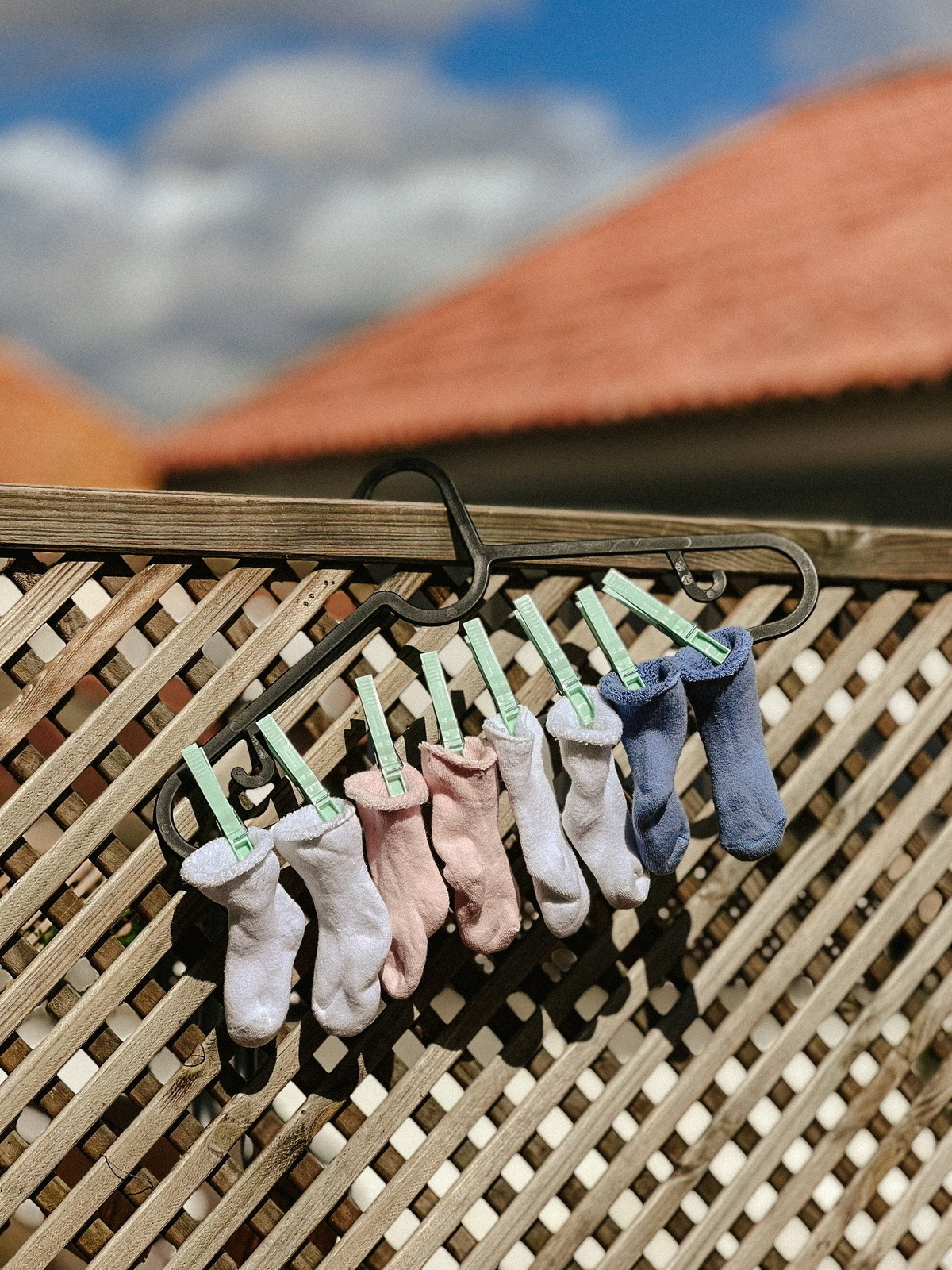 Baby socks in various colors are clipped with green clothes pins on a black hanger, drying on a wooden lattice against a backdrop of a blue sky with clouds and rooftops.