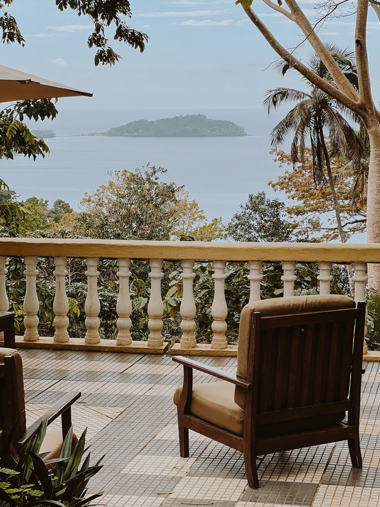 A serene patio scene overlooking the ocean with an island in the distance. Two wooden chairs with beige cushions are positioned on a tiled terrace, surrounded by trees and vegetation. A stone balustrade separates the patio from the lush greenery below.