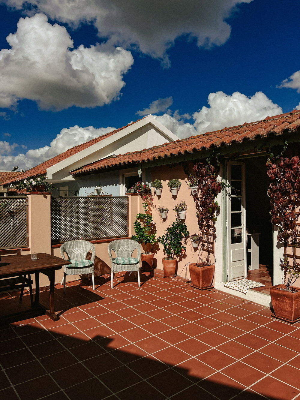 Terracotta-tiled patio with wicker chairs and a wooden table. Potted plants and climbing vines decorate the pink stucco wall. A white door opens to the interior. The background features a tiled roof under a bright blue sky with fluffy clouds.