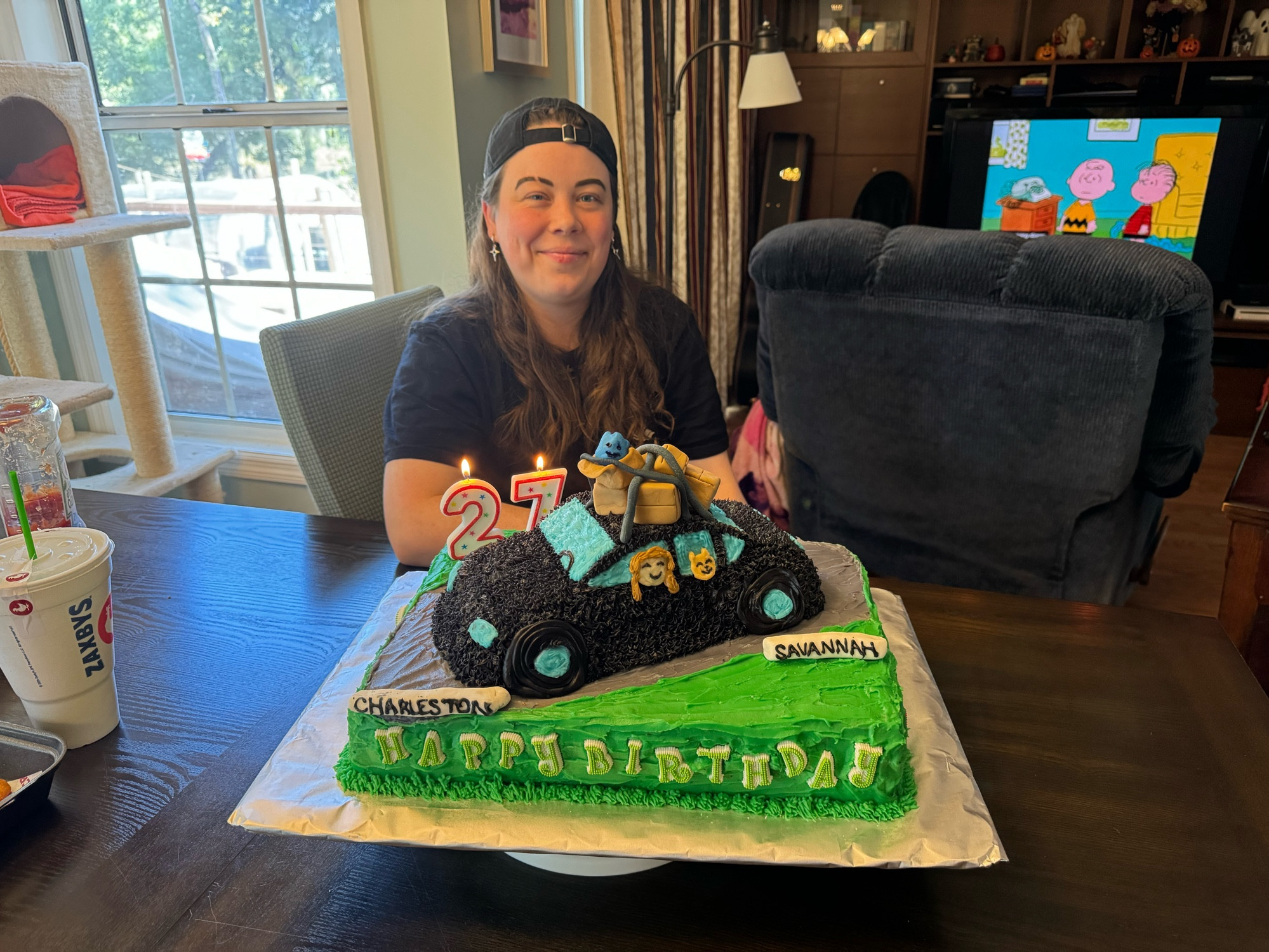 Me sitting in front of my birthday cake. It has a sheet cake bottom and a car-shaped top layer with fondant boxes and straps on top. A crude sonic-shaped fondant plushie is in one of the open boxes. My head and my cat’s head are visible in the side windows. Signs at the front and back of the cake read “Charleston” and “Savannah”. Candy pieces along the bottom read “Happy Birthday” and two lit “27” candles. 