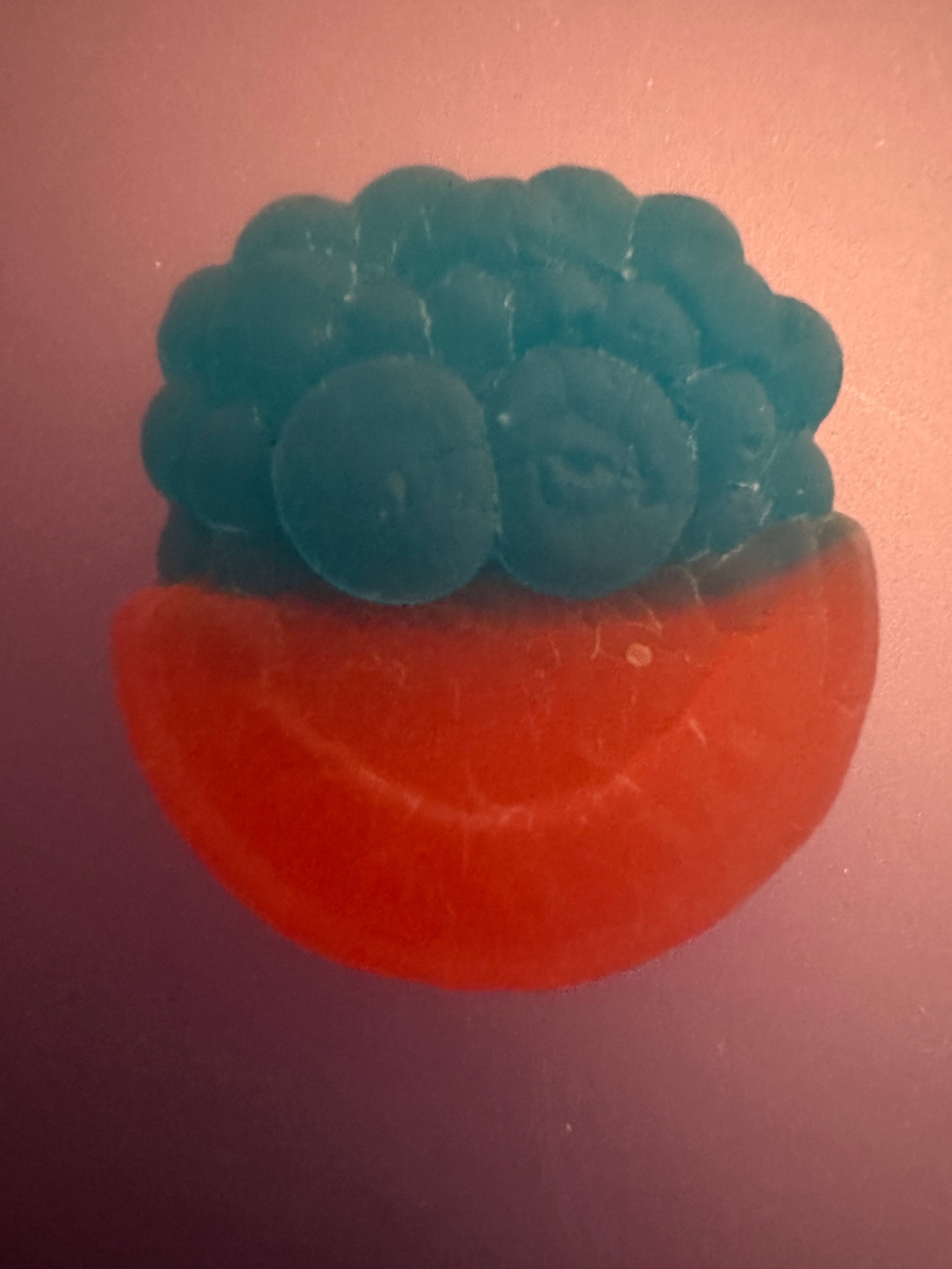 a picture of a piece of candy. the top is “blue raspberry” and has been designed to look like a raspberry. two large drupelets represent eyes. the bottom is red (pink actually but the lighting makes it inaccurate) and is supposed to be a watermelon section. the rind and some of the flesh represent a smiling mouth. watermelon seeds represent teeth, although these are not very visible in the picture. overall i find it horrifying