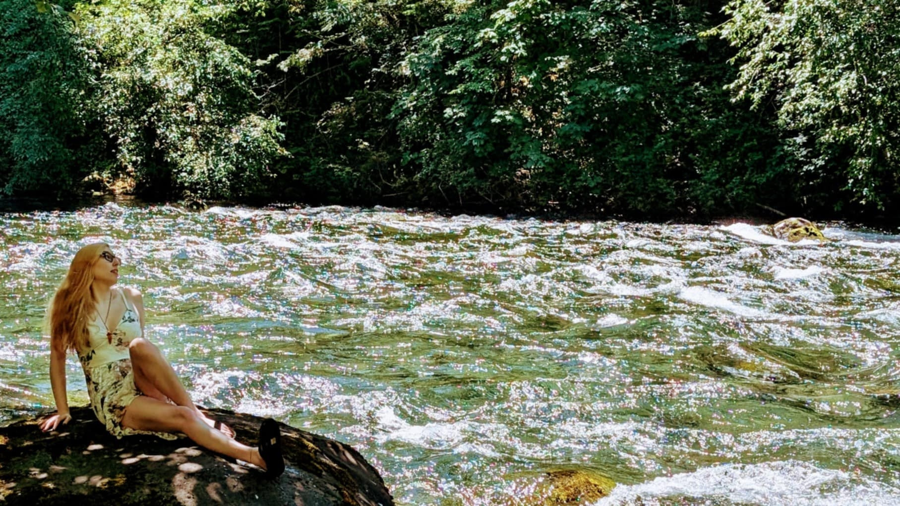 Blonde woman wearing a flowery dress by the river, posing up a *storm* while laying on a rock