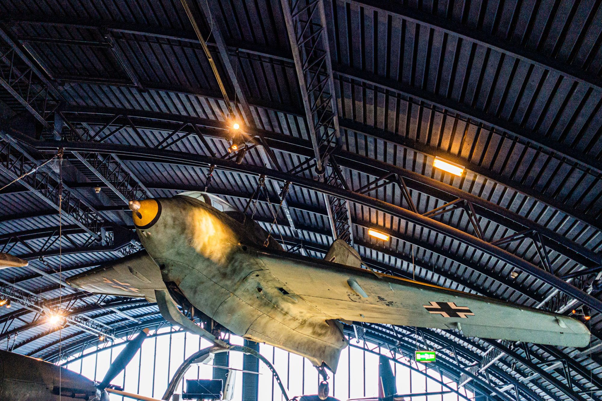 This photo features a vintage German aircraft from World War II, suspended from the ceiling of a museum. It has a unique, delta-wing design and is painted in a camouflage pattern. The nose is pointed and features a large, yellow radome. The aircraft is suspended from cables attached to its wings and fuselage. The background shows the museum's ceiling with exposed metal beams and lighting.