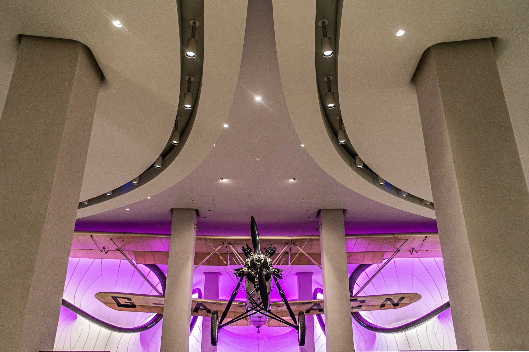 A grand, symmetrical interior space with a high, curved ceiling featuring soft, indirect lighting. Two rows of thick, cylindrical columns flank the centre of the room. Suspended from the ceiling is a vintage biplane, its wings and propeller highlighted by a dramatic purple light. The background is a neutral, off-white colour. The overall atmosphere is one of grandeur and historical significance, suggesting a museum setting.