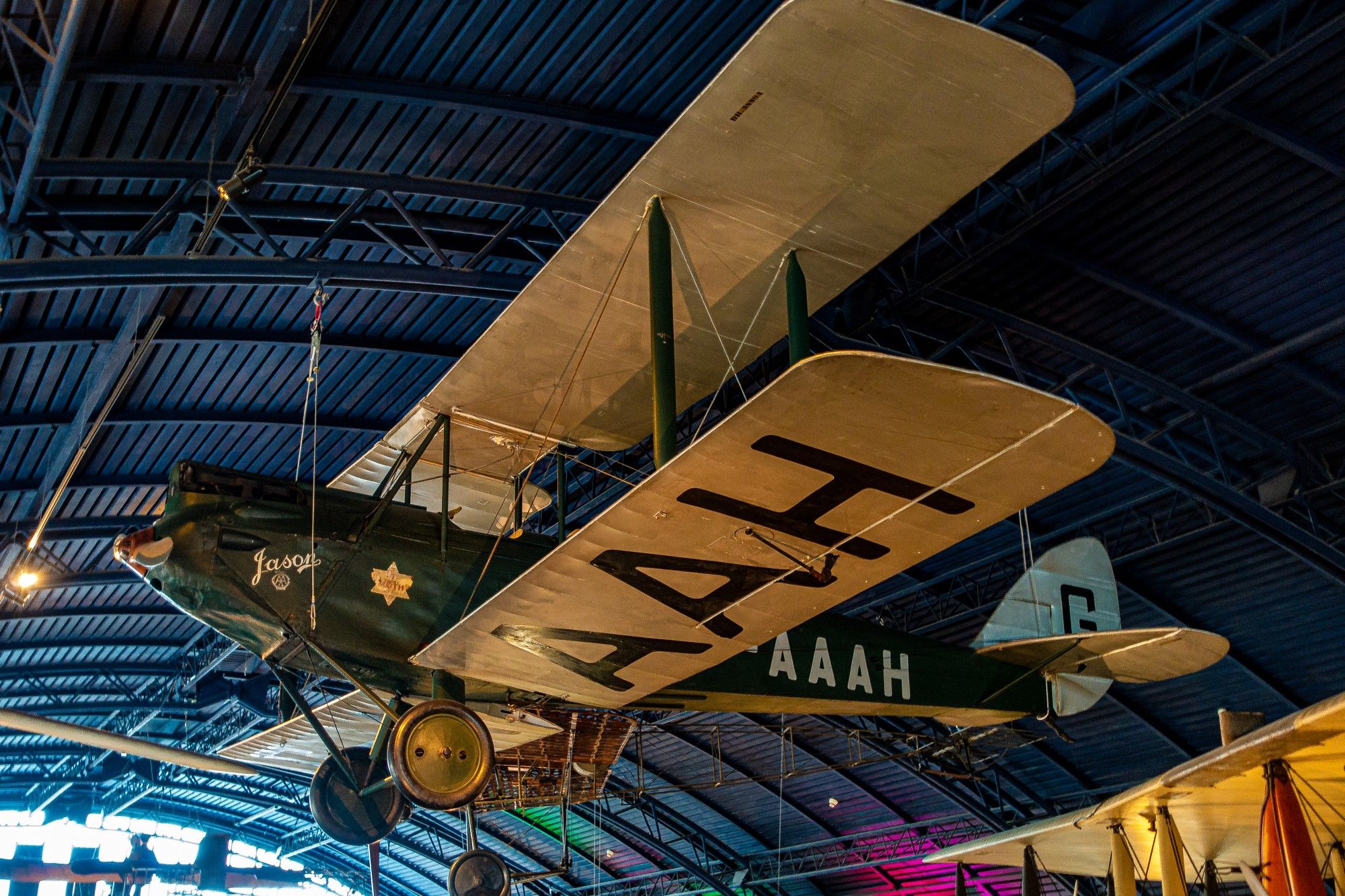 This photo features a vintage biplane aircraft hanging from the ceiling of a museum. The aircraft is primarily white with dark green markings. The wings are large and have visible struts and wires. The nose of the plane is rounded and has a prominent propeller. The registration number "G-AAAH" is painted on the side of the fuselage. The background shows the ceiling of the museum with exposed metal beams and lighting fixtures.