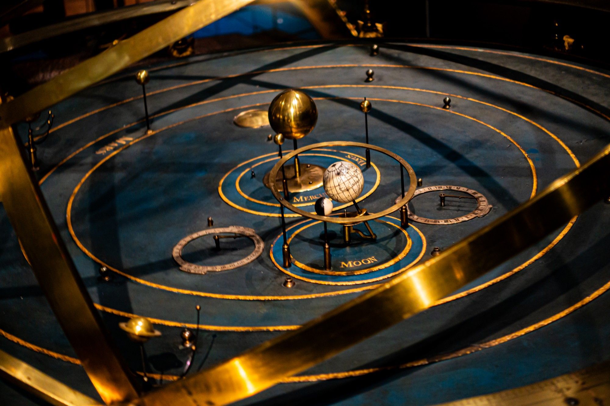 This photo shows a historical orrery, a mechanical model of the solar system. It consists of a circular base with concentric rings representing the orbits of the planets. A central sphere represents the Sun, and smaller spheres on rods represent the planets. The planets are labelled with their names, including Mercury and Moon. The model is made of brass and has a dark blue background. The overall appearance is antique and intricate.