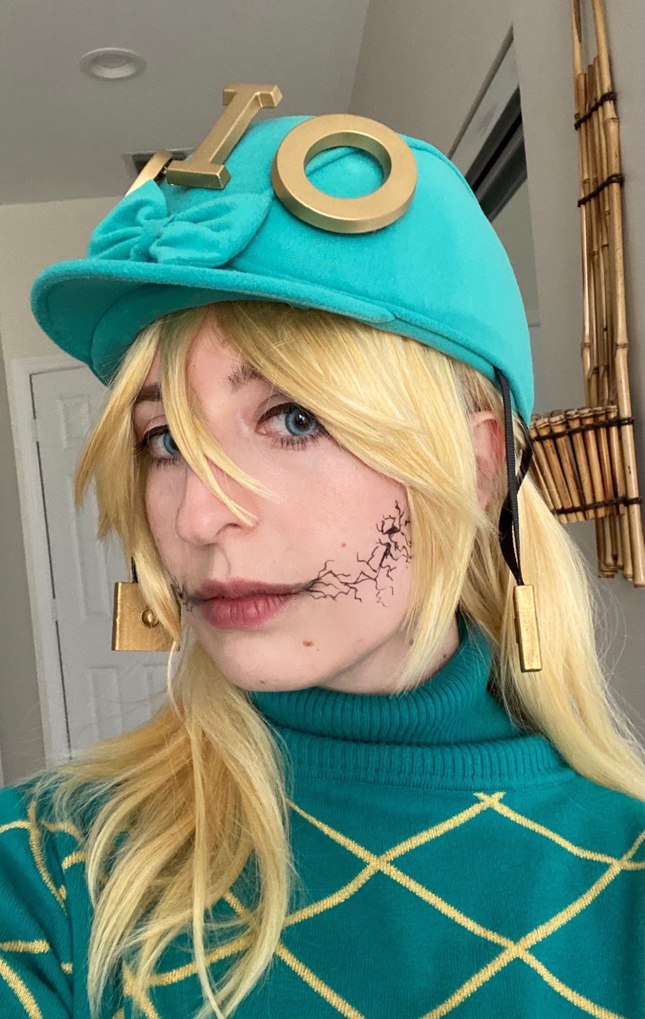 Mar is dressed as Diego Brando from Jojo's Bizarre Adventure part 7: Steel Ball Run in this selfie. She wears a teal turtleneck jumper with a yellow grid pattern and a teal riding helmet with large gold letters that spell out "DIO". She has yellow blonde shoulder-length hair. A skin cracking effect extends from the corner of her lips.