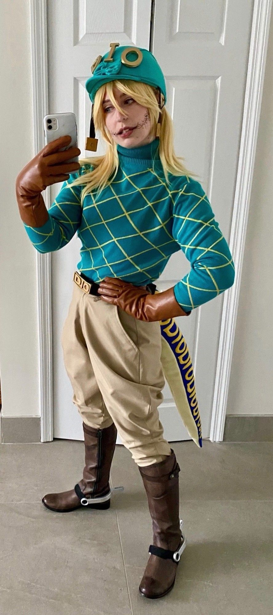 Mar is dressed as Diego Brando from Jojo's Bizarre Adventure part 7: Steel Ball Run. She wears a teal turtleneck jumper with a yellow grid pattern, saddle brown gloves, beige jodhpurs, brown riding boots, silver spurs, a belt with a gold buckle reading "DIO", and a teal riding helmet with large gold letters that spell out "DIO". She has yellow blonde shoulder-length hair, and a blue and beige velociraptor tail with yellow letters repeatedly spelling out "DIO". A skin cracking effect extends from the corner of her lips.