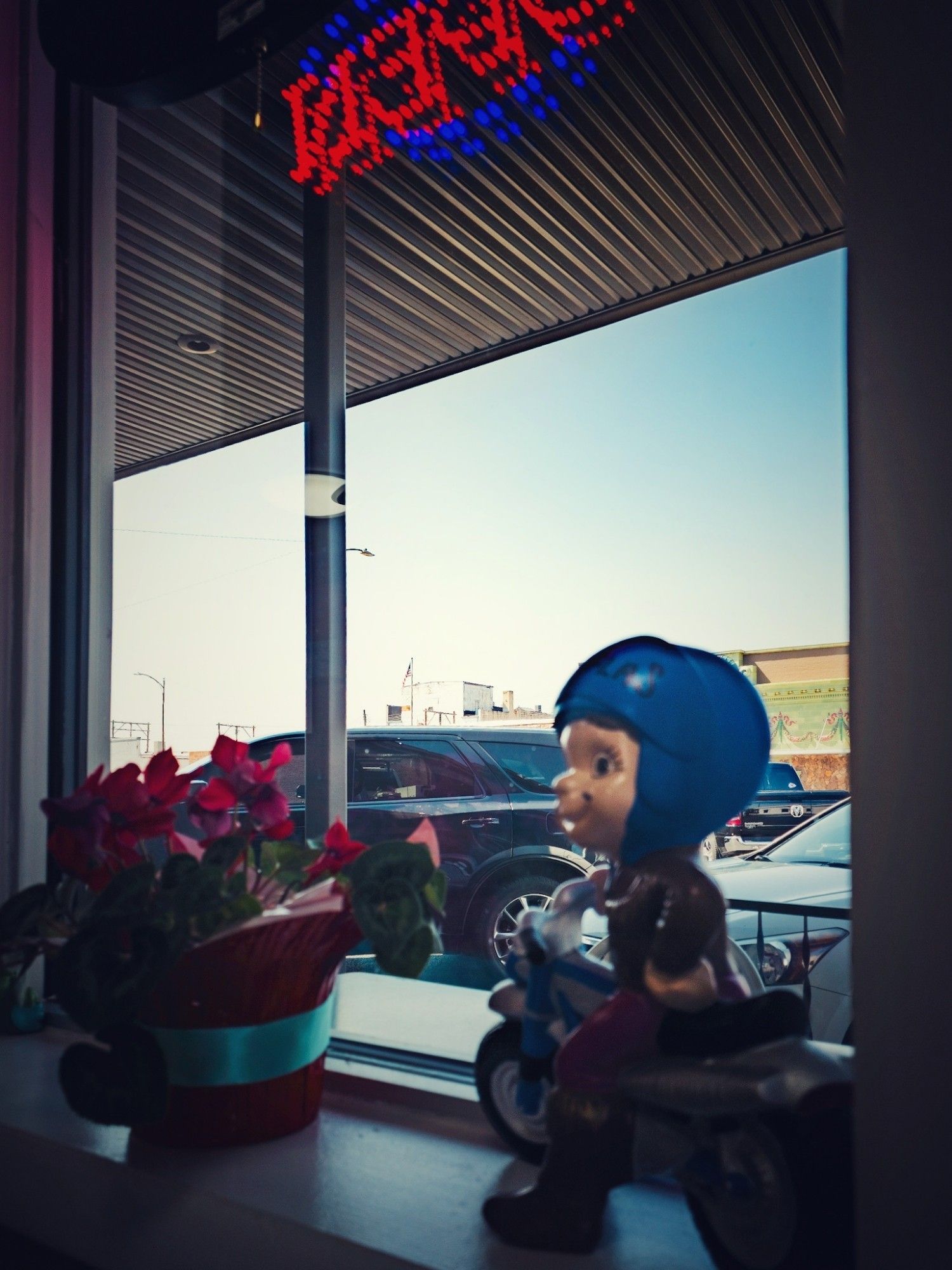 A view through the window of diner. On the window ledge is a flowering plant and a toy figurine of a boy on a motor bike. Overhead is the electric open sign reflecting on the metal awning. Barely visible beyond the parked cars is the green and pink ornate facade of a defunct theatre and a weathered white industrial building.