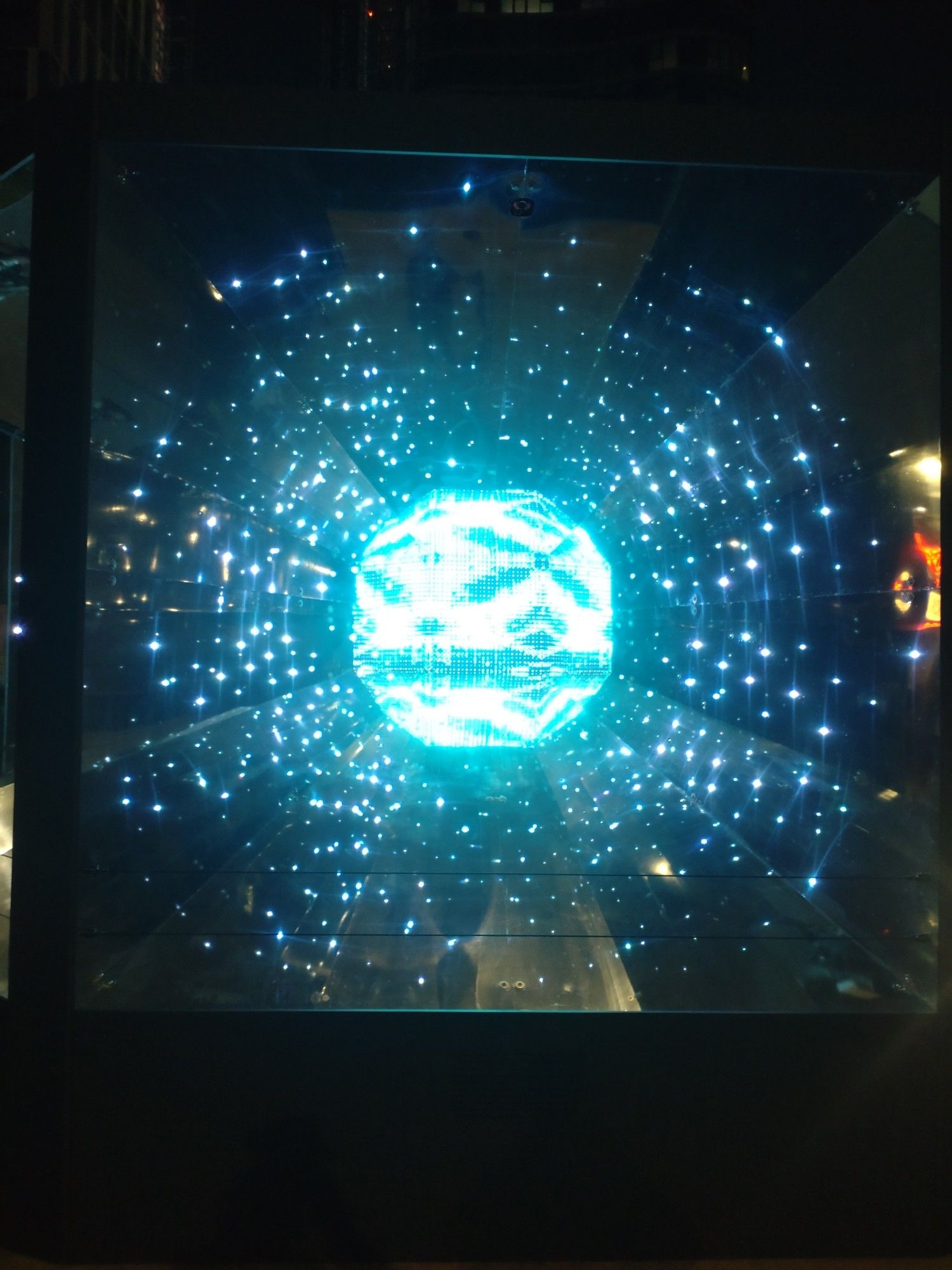 A glass box with a large ball of light in the centre that changes in colour and intensity. Surrounding smaller individual lights change, spreading outwards in the cube