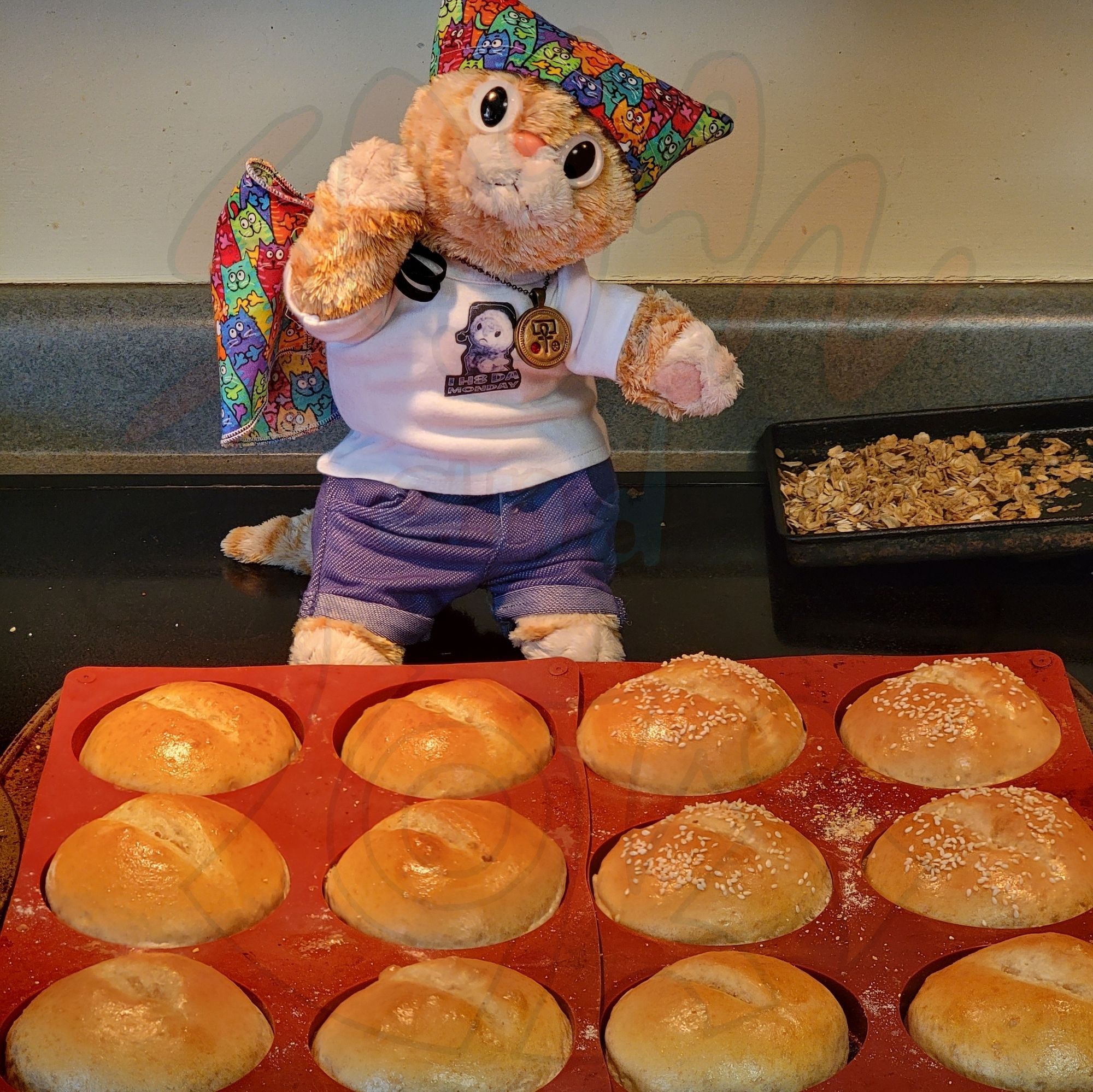 An orange plush cat in jeans and a tee wearing a colorful bandana and holding a matching apron stands behind silicone bun trays with 8 plain and 4 sesame shiny, golden rolls.