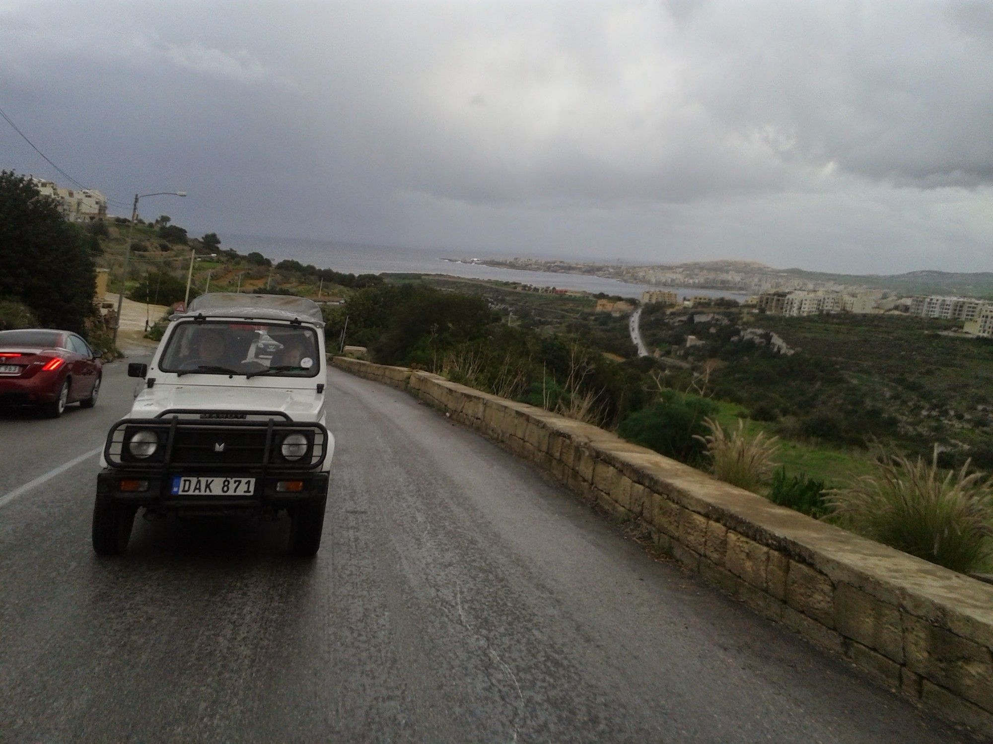 Jeep-Fahrt auf Malta,  im Regen. Hinter uns ein Jeep,  ein Grünstreifen rechts der Straße. Im Hintergrund das Meer und dunkle Wolken am Himmel.