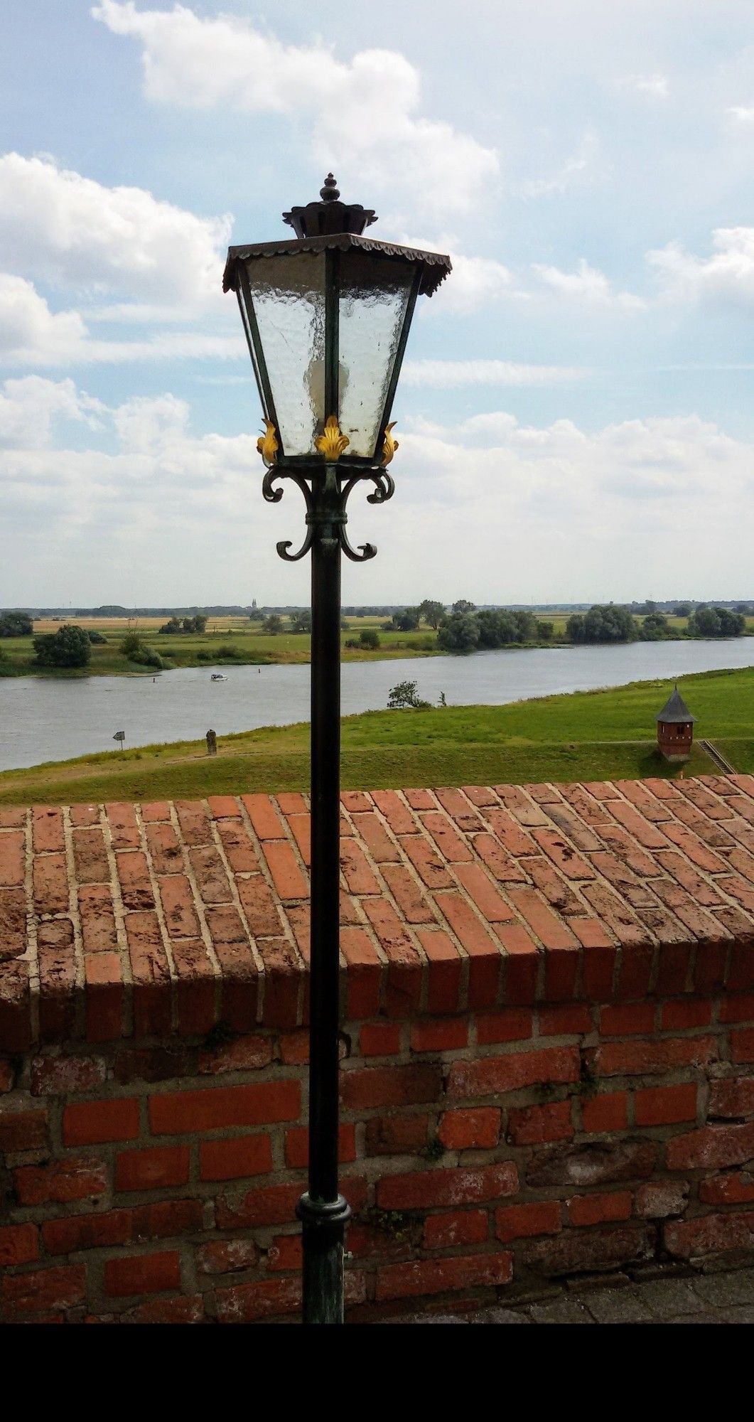 Von einer roten Backsteinmauer auf eine alte Laterne blickend, liegt unterhalb und von Wiesen gesäumt der kleine Bach , die "Elbe".  Der Himmel ist teils himmelblau mit weißen Wolken gespickt.