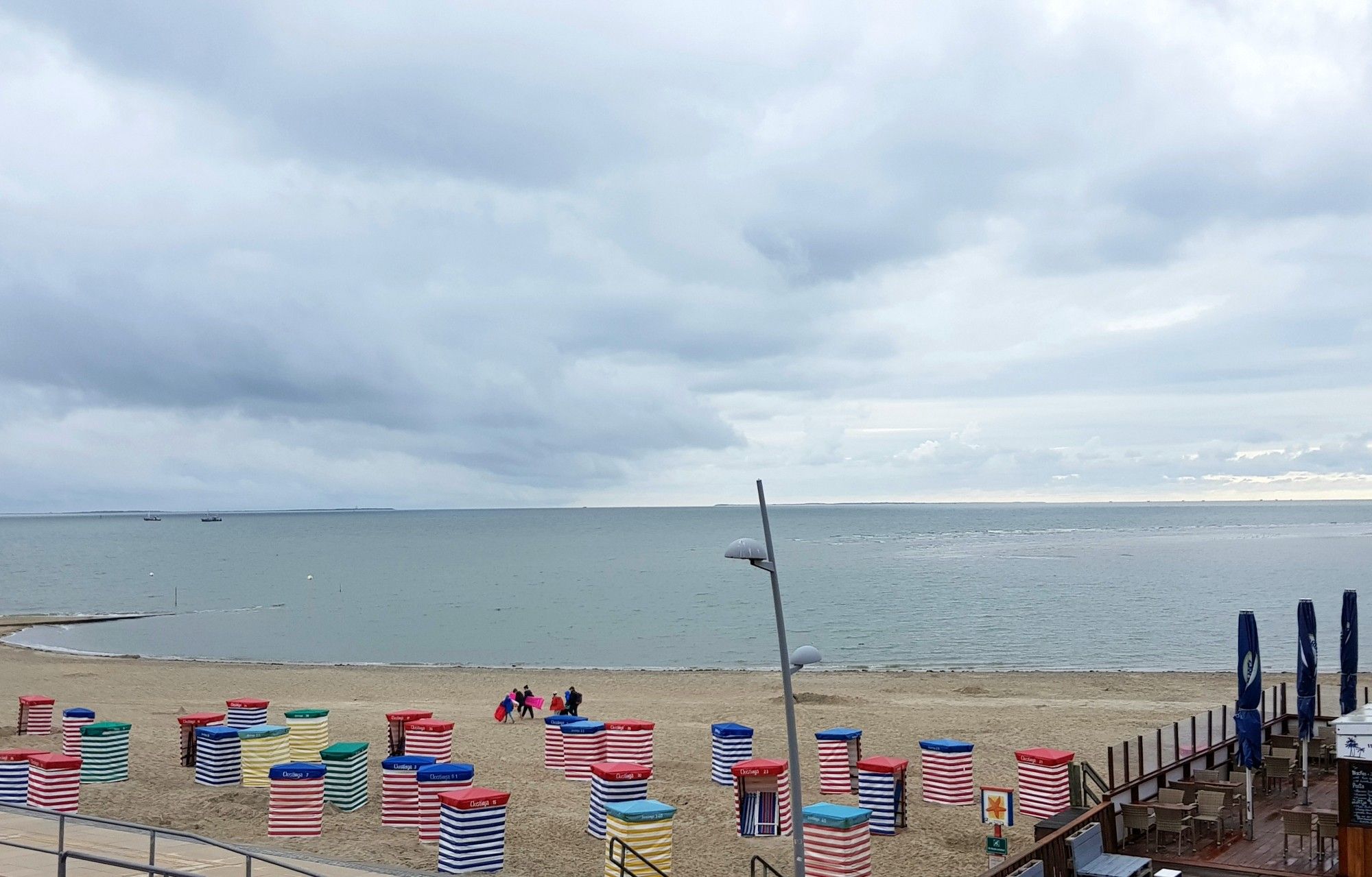 Von der Promenade auf den Strand mit bunten Strandkörben geschaut. Rechts eine Lokalität. Und der Blick über die glatte Nordsee gerichtet. Der Himmel ist bewölkt.