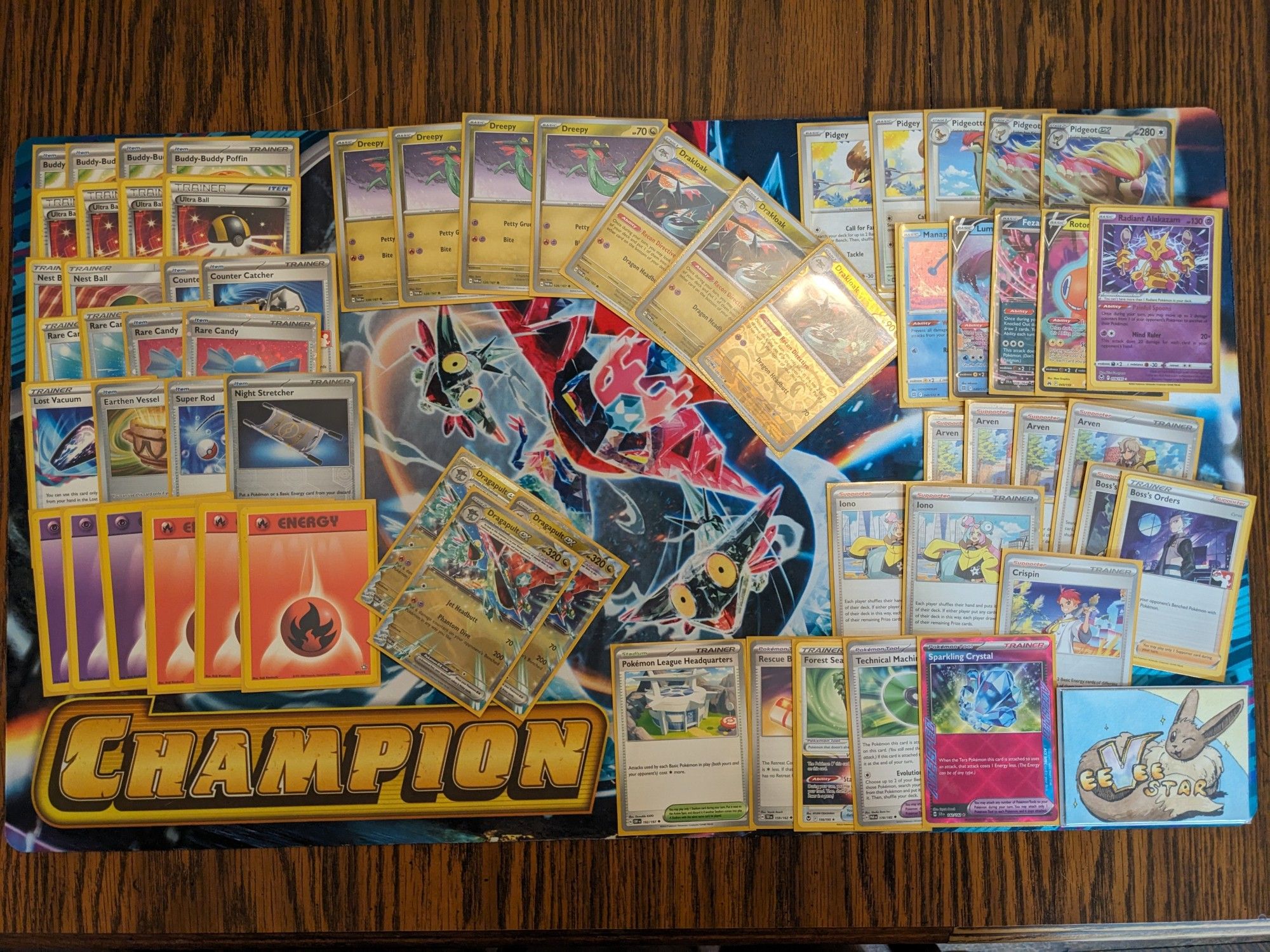 A Pokemon TCG deck spread out over a playmat that says "champion" in the corner. The deck is based on Dragapult cards, which coincidentally is also the Pokemon depicted on the playmat. There's also a cute watercolor painted "eevee-star" marker token in the corner.