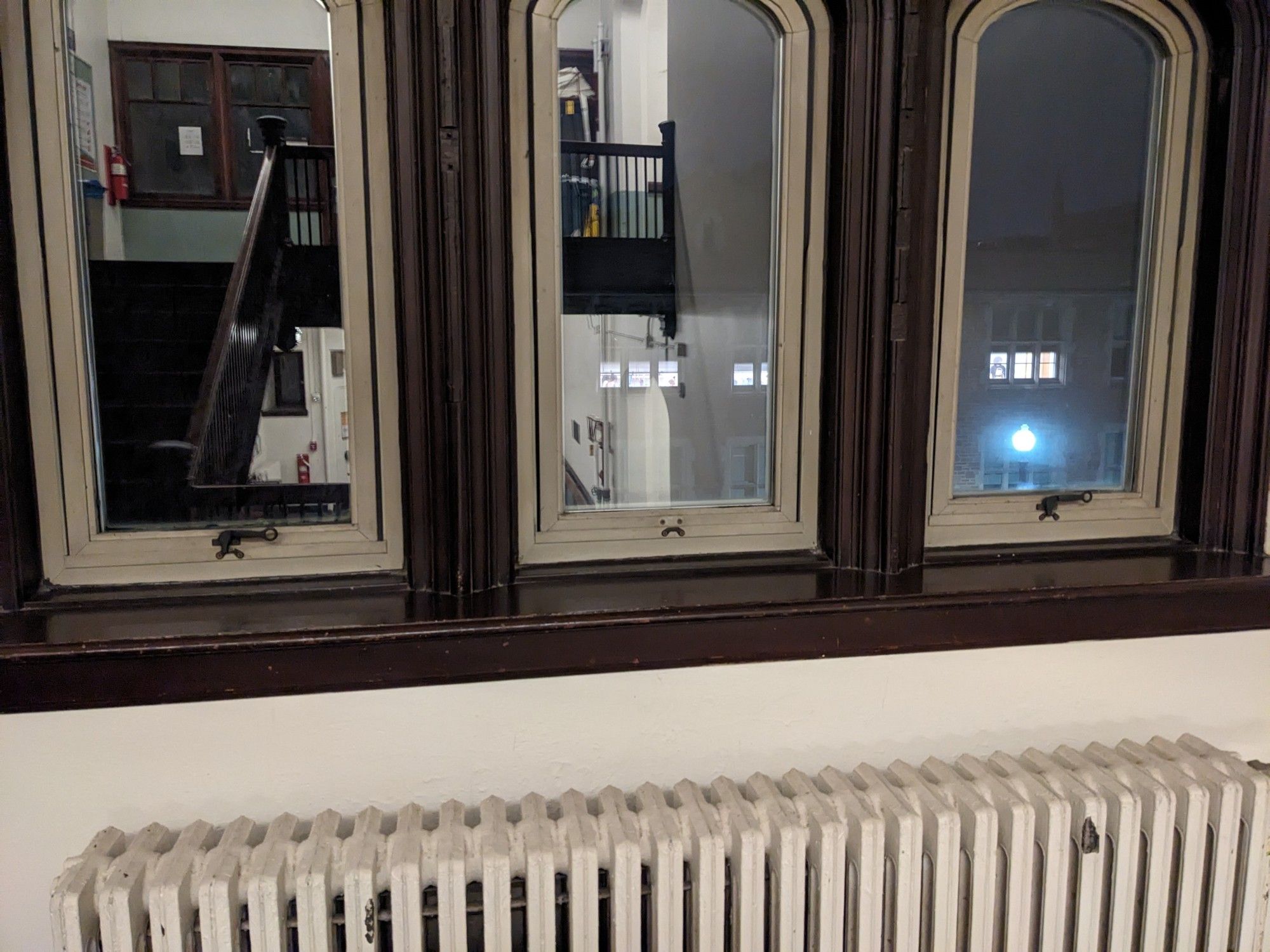 Three windows in a row in an old building. Two of them have devices at the bottom on the frames for locking them closed. On the middle one, most of the device is missing, leaving some hardware in the shape of a frowny face.