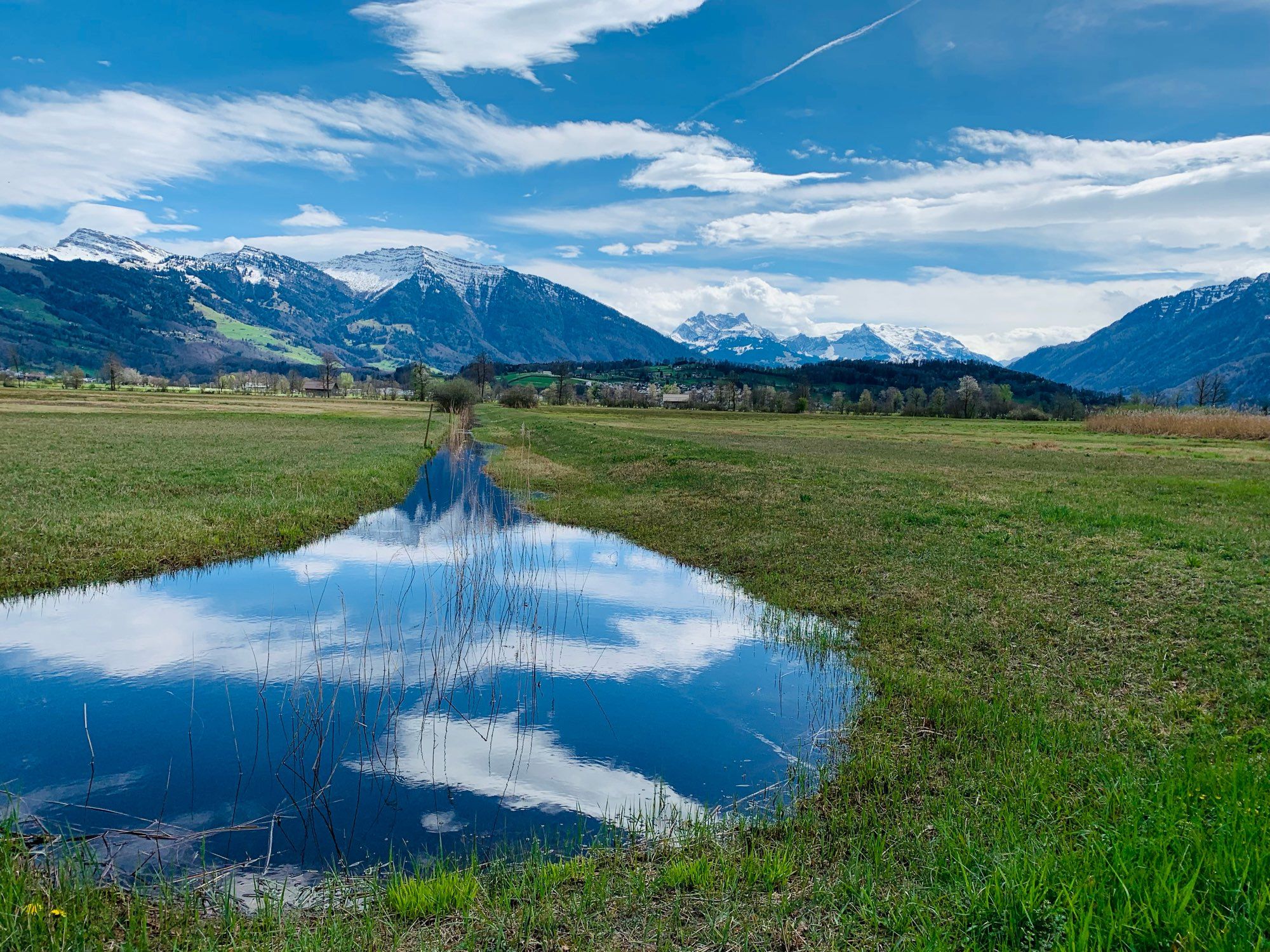 Ein kleiner Teich auf einer grünen Wiese. Der blaue Himmel und die weissen Wolken spiegeln sich im Wasser. Dahinter schneebedeckte Berge und Wald.