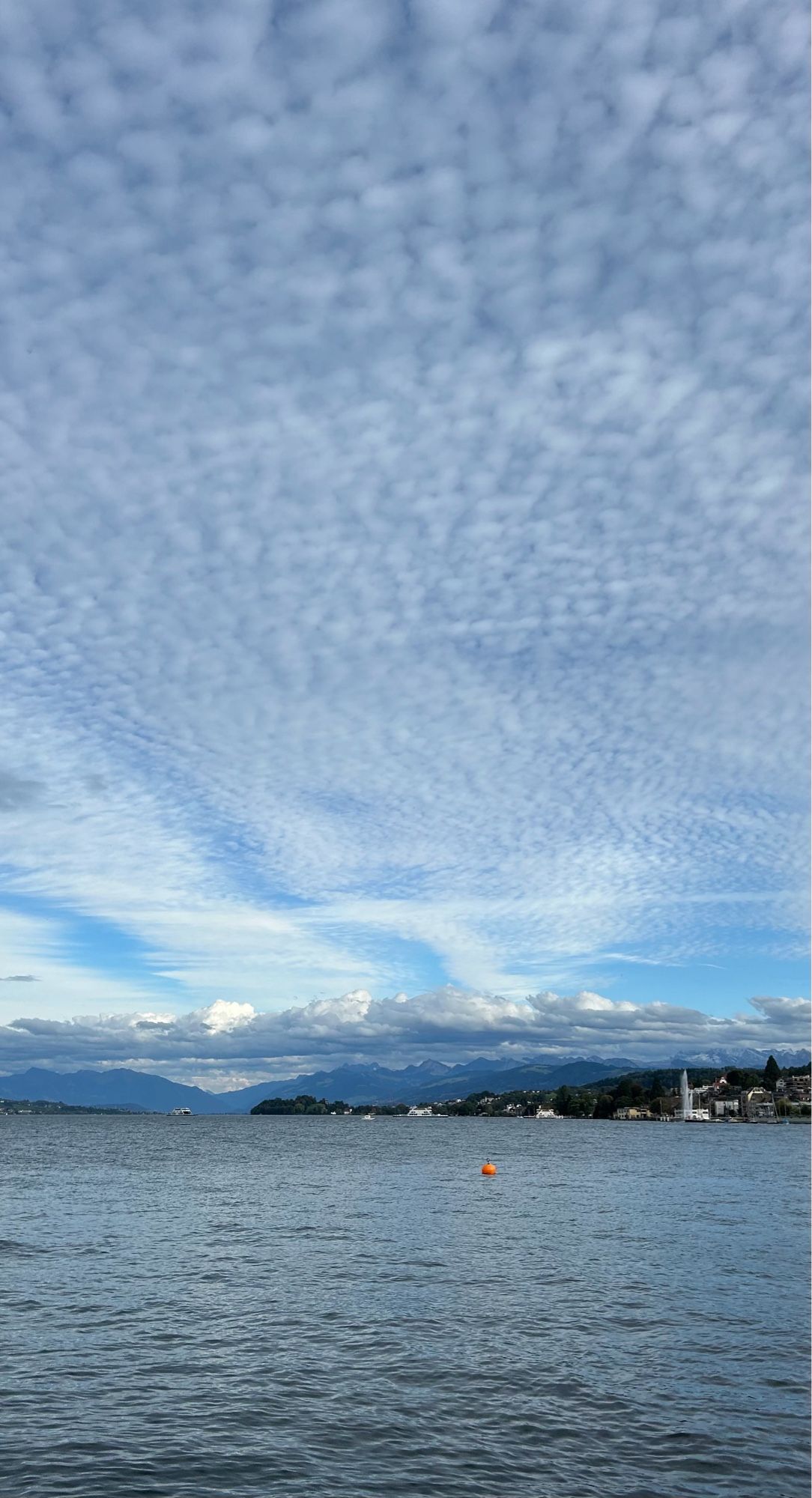 A view over the lake with mountains in the distance. The majority of the picture is dominated by the sky, featuring distinctive, striking cloud formations.