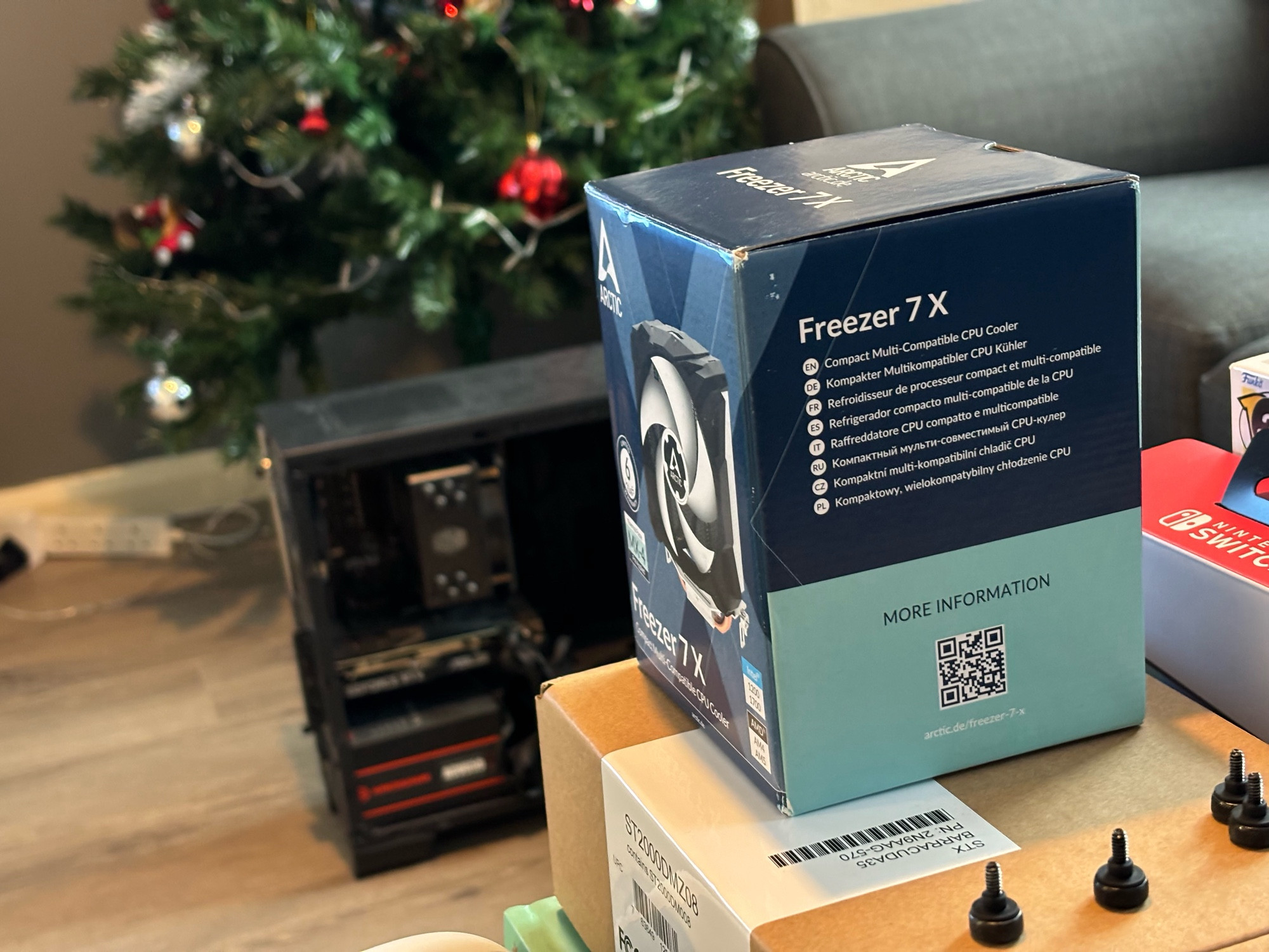 A photo of a new CPU fan in box with some screws near it and a PC in the background in front of my Christmas tree