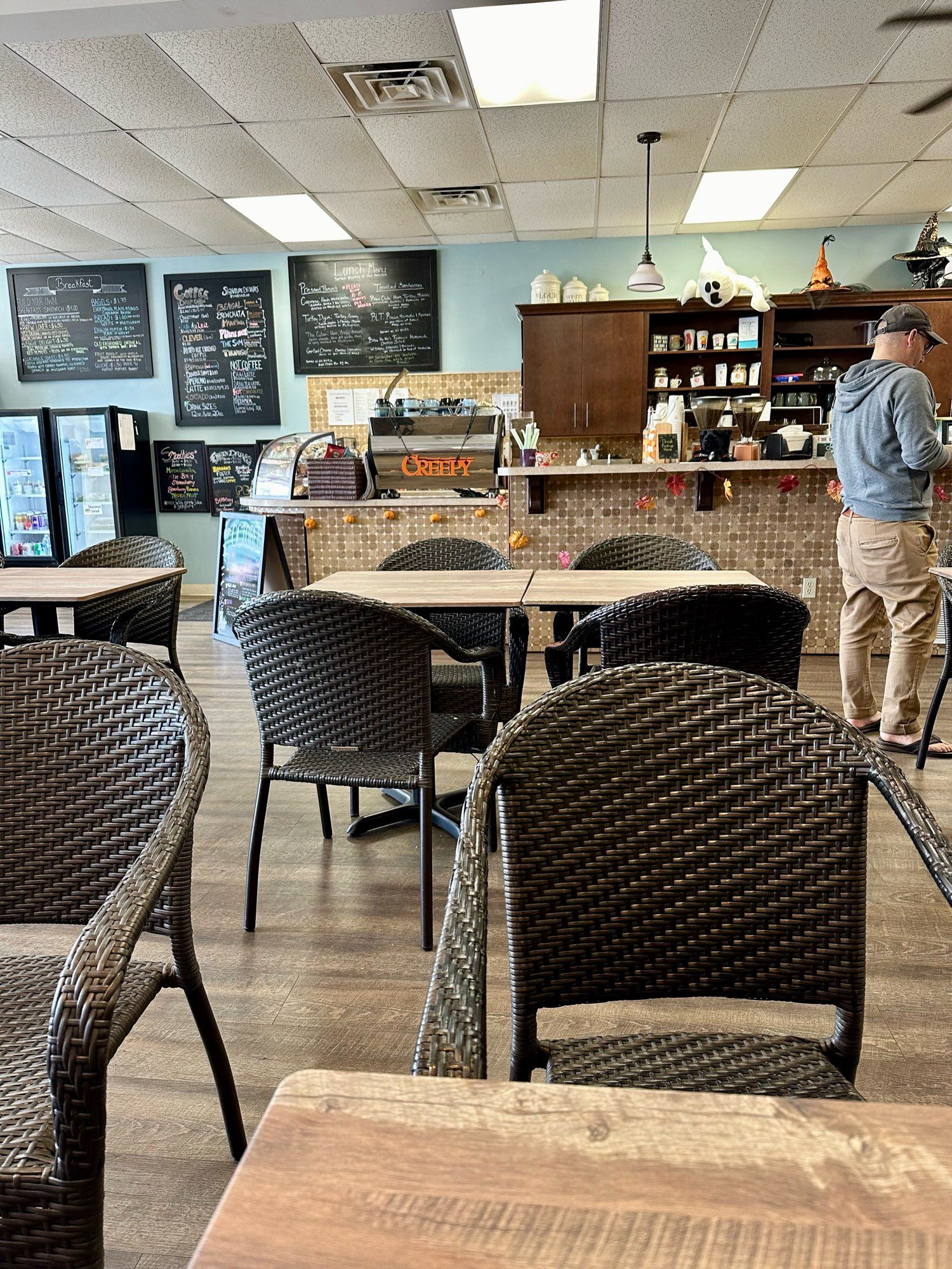 Counter of a coffee shop from the opposite wall with small, square wooden tables and wicker looking chairs arrayed in front of it. Handwritten signs are visible on the wall and an orange Halloween decoration that says “CREEPY” is hung on the back of the espresso machine.