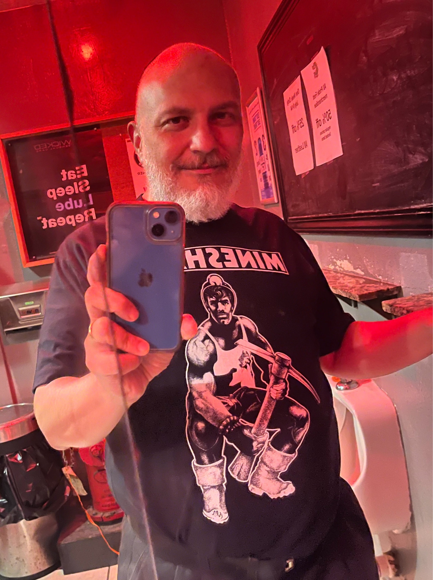 Man poses in front of a mirror. He has a retro Mineshaft bar t- shirt on.