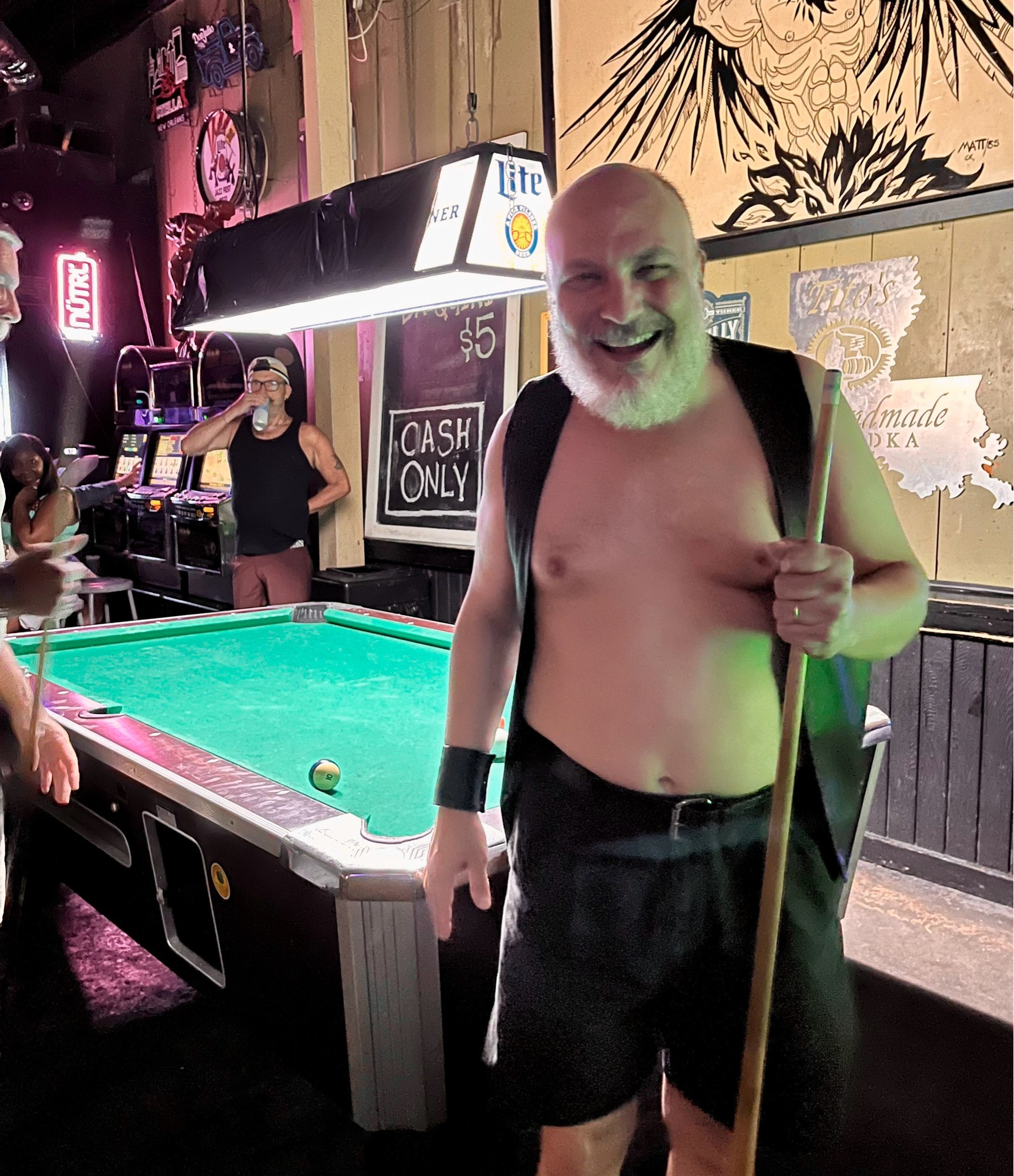 A man stands in front of a pool table holding a cue stick. He is wearing a leather bar vest, no shirt, and shorts.