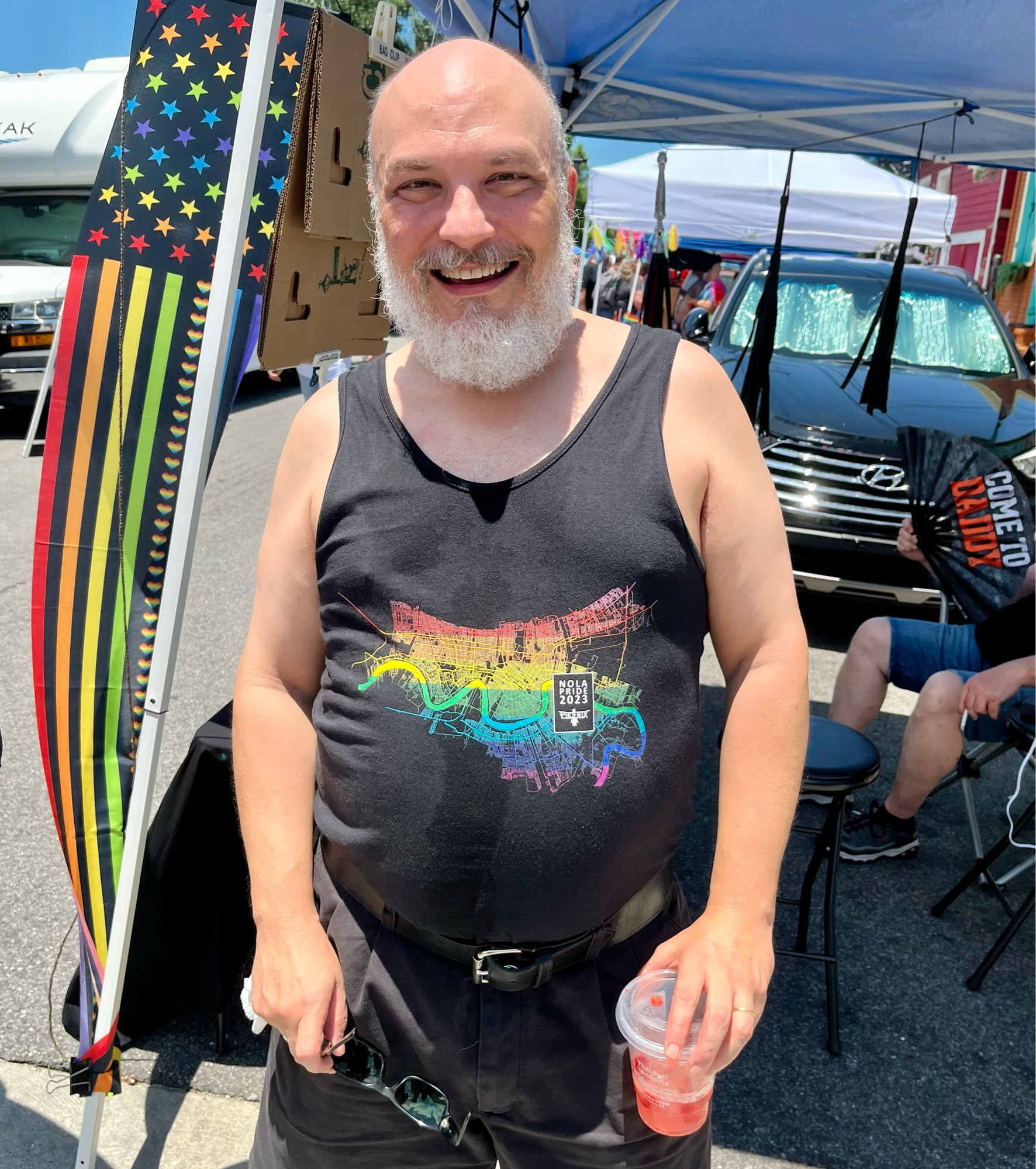 A man with a white beard and close cropped hair stands in sunlight wearing a pridefest shirt that depicts the city of New Orleans in rainbow colors